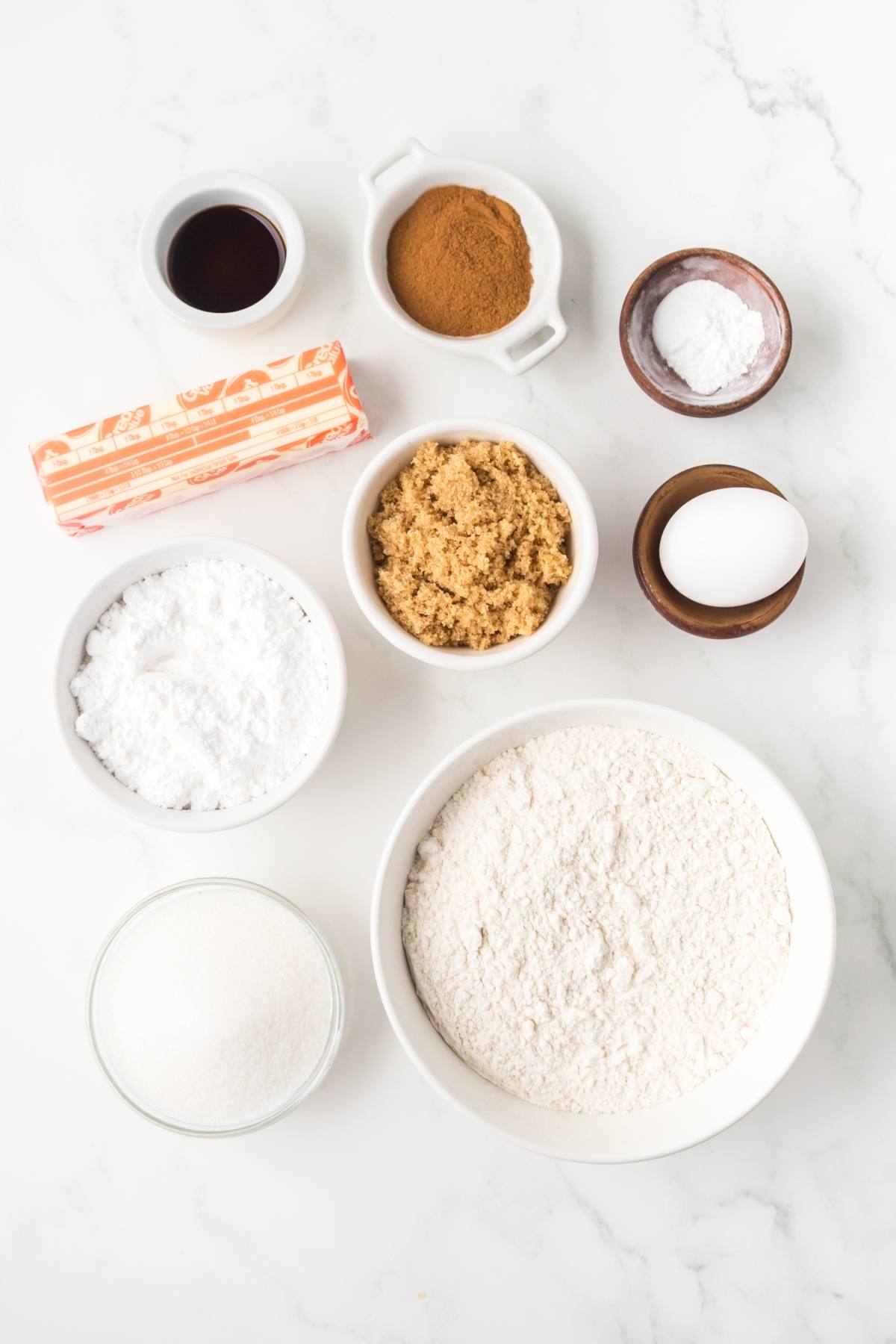 Baking ingredients on a white counter: flour, egg, sugars, butter, vanilla, cinnamon, baking soda, and powder.