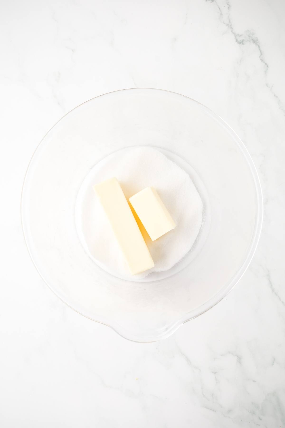 A glass bowl with sugar and two sticks of butter on a white marble surface.