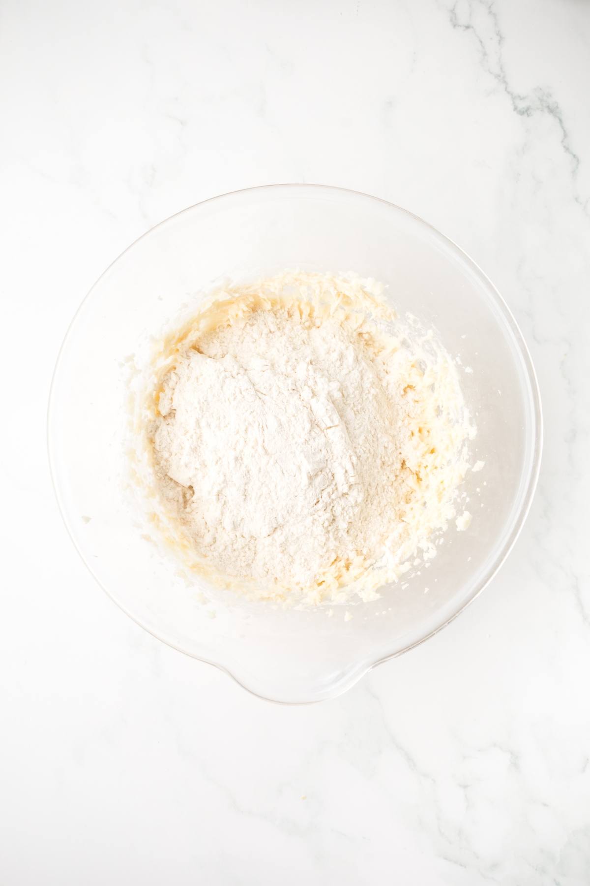 A glass mixing bowl with flour and creamed ingredients on a white marble surface.