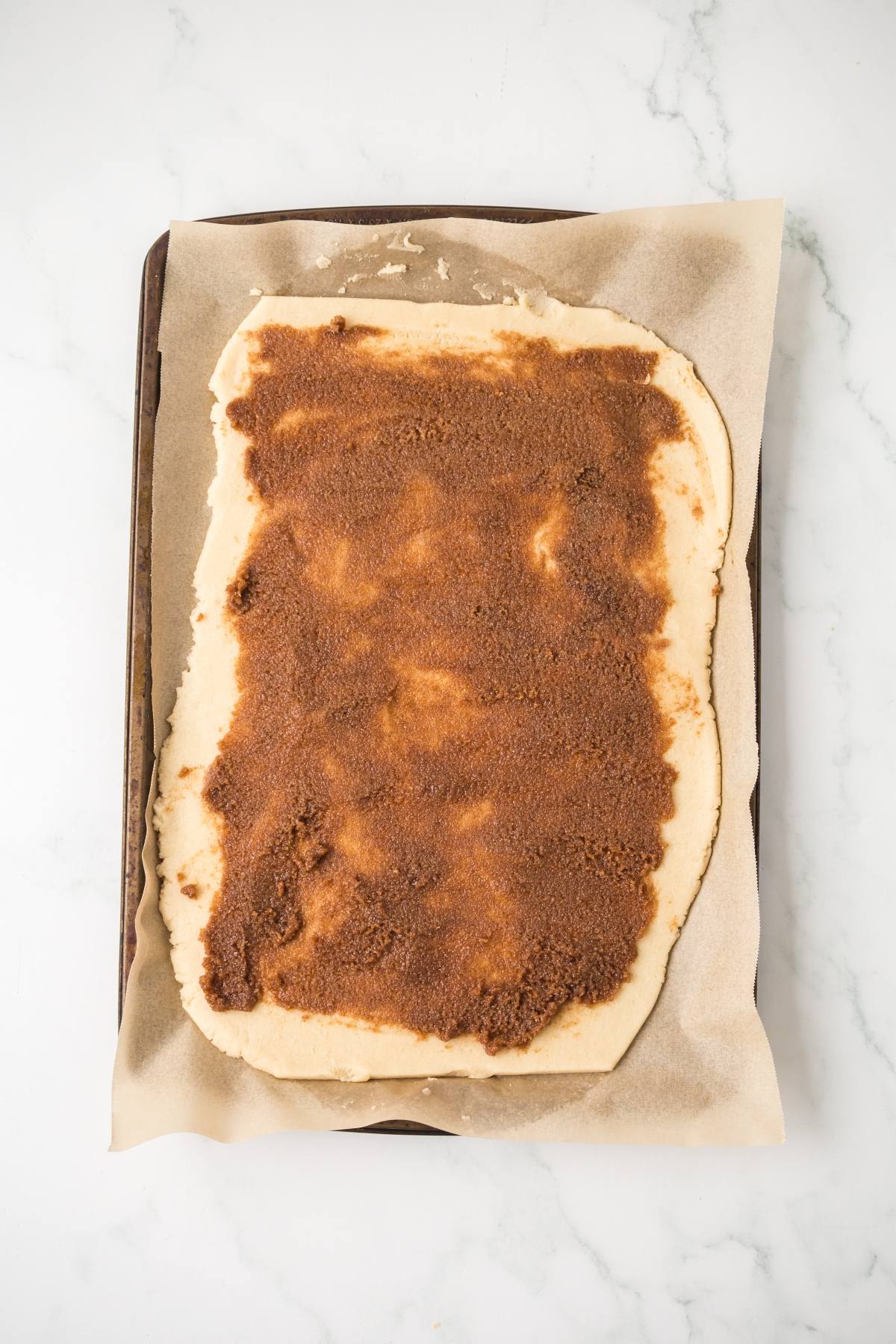 Rectangular dough spread with cinnamon and brown sugar mixture on parchment paper, ready to bake.
