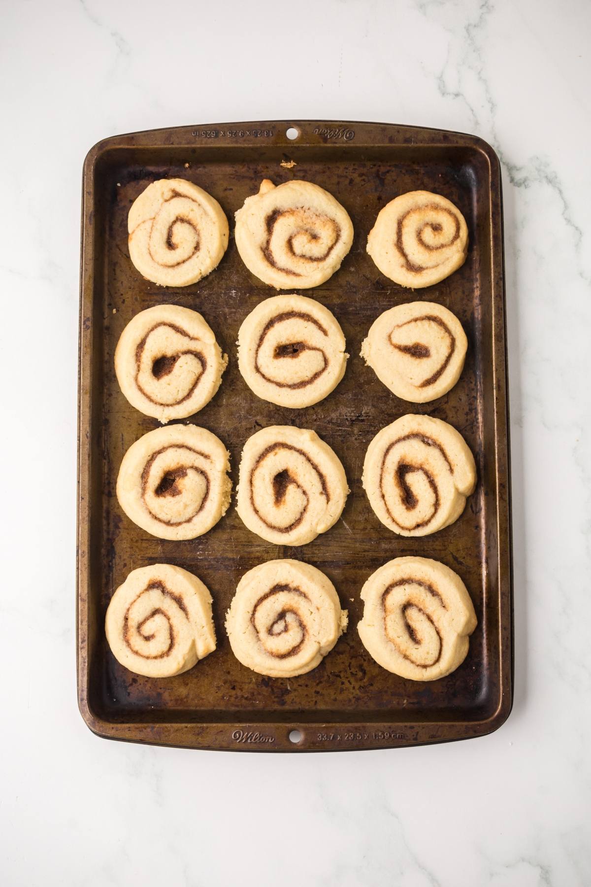 A baking tray with twelve unbaked cinnamon rolls arranged in rows on a white marble surface.