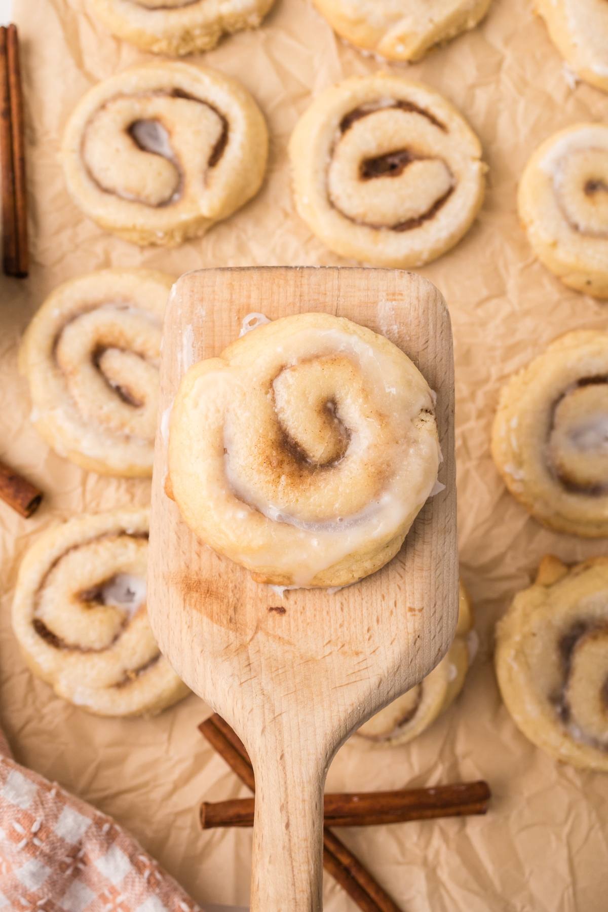 A wooden spatula lifts a glazed cinnamon roll surrounded by more rolls on parchment paper.