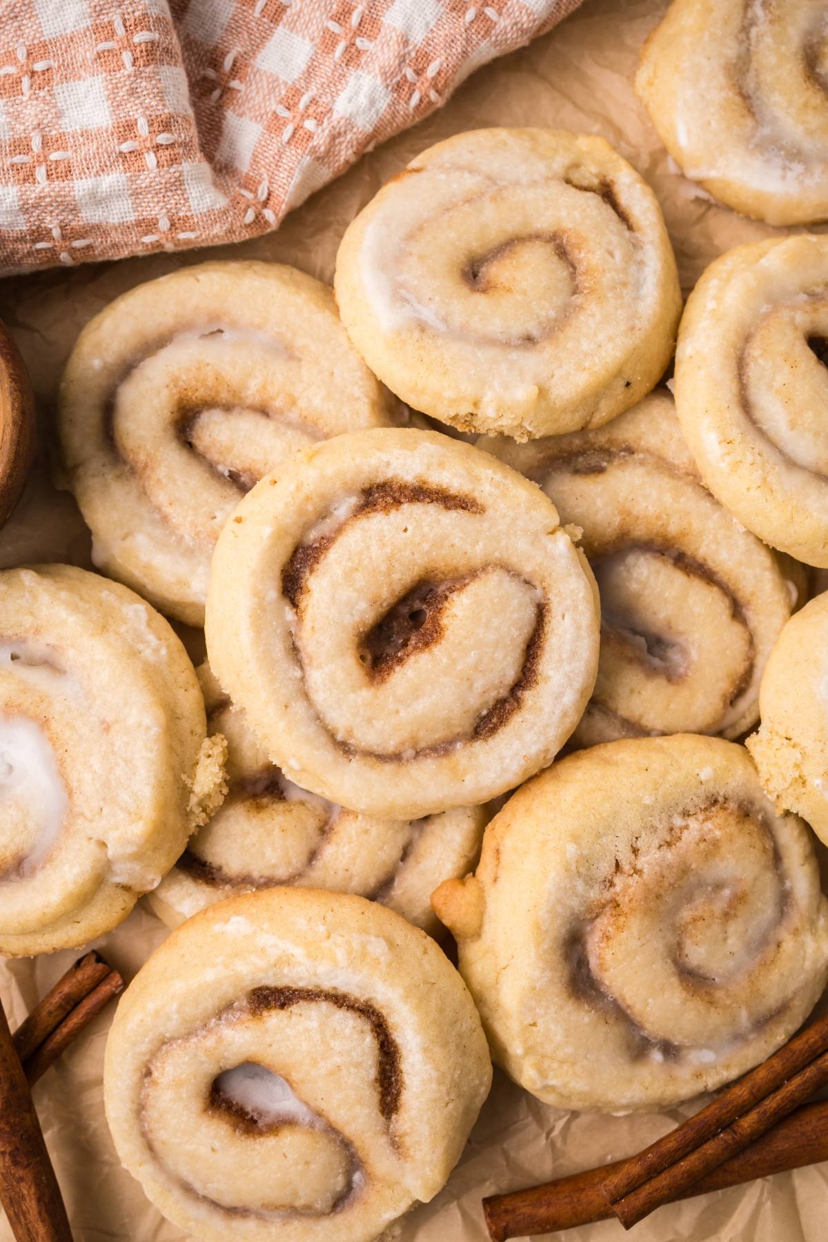 A pile of cinnamon swirl cookies on parchment paper, with a checkered cloth and cinnamon sticks beside them.