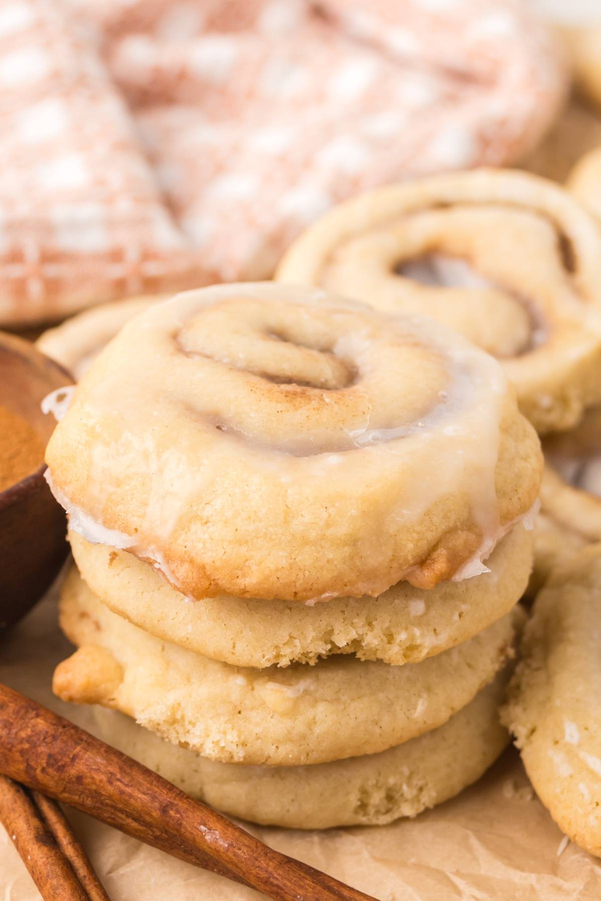 Stack of glazed cinnamon roll cookies with cinnamon sticks and a checkered cloth in the background.