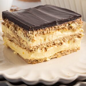 A slice of Eclair Cake with chocolate topping and creamy filling rests on a stack of white plates.