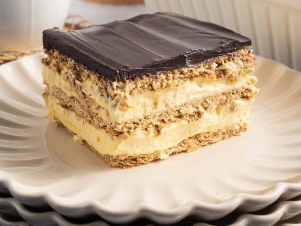A slice of Eclair Cake with chocolate topping and creamy filling rests on a stack of white plates.