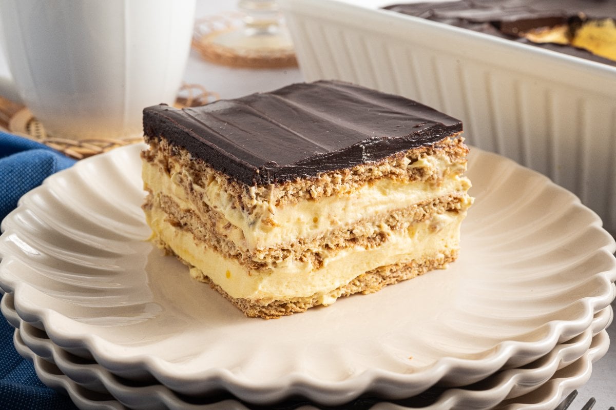 A slice of layered Eclair Cake sits on a cream-colored plate, topped with rich chocolate and filled with smooth, creamy filling.