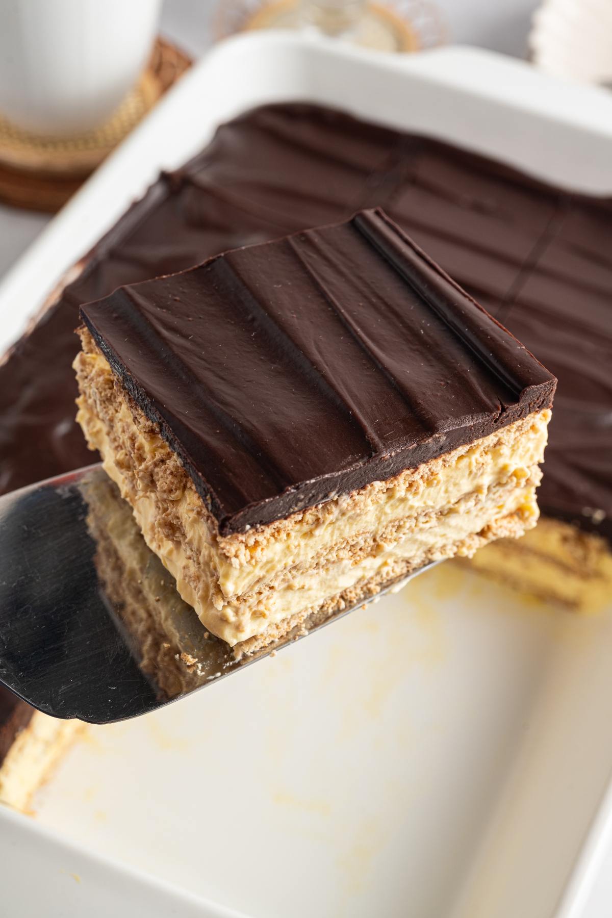 A spatula lifts a slice of layered chocolate and cream dessert from a white dish.