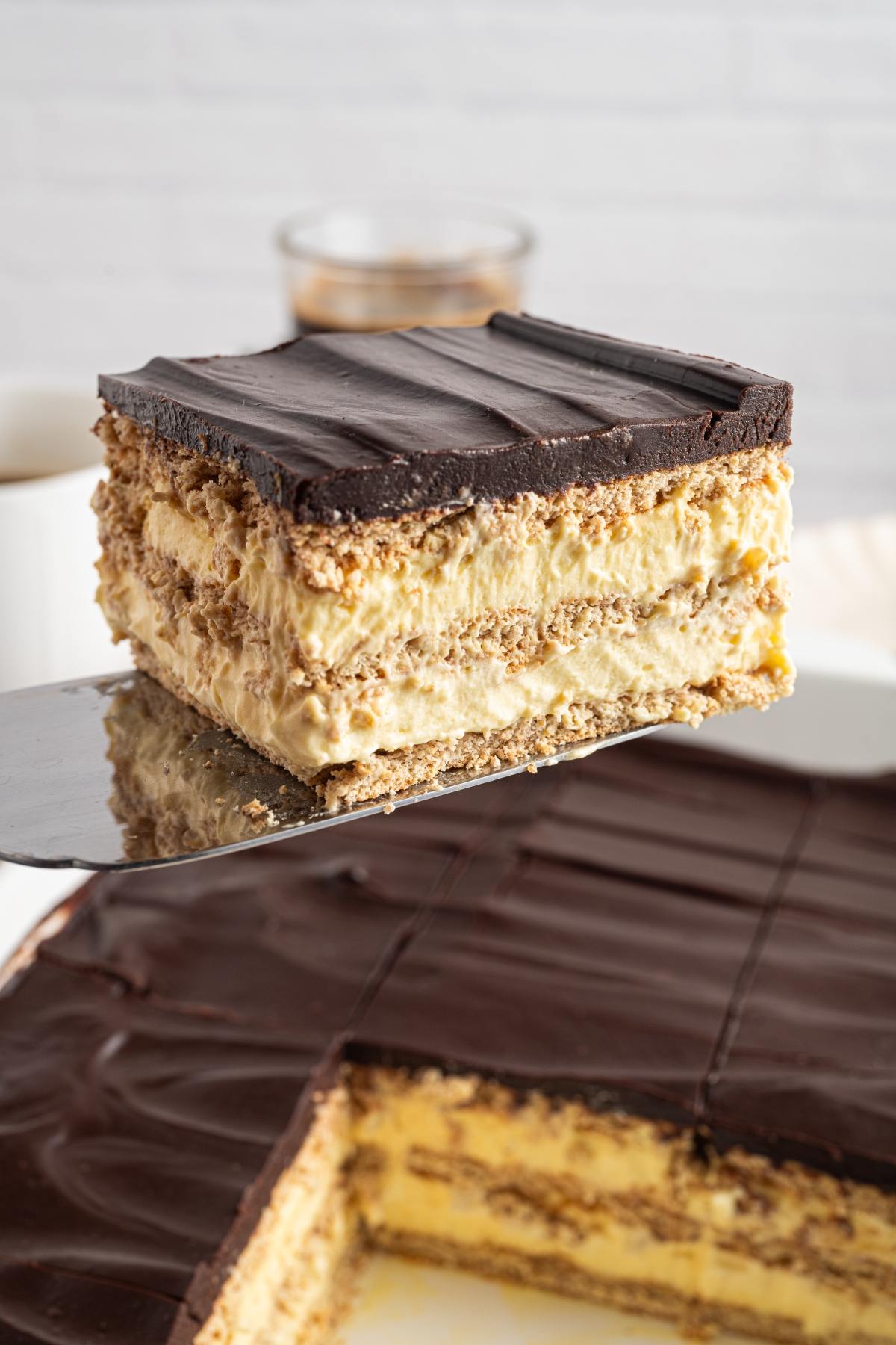 A slice of layered chocolate-topped dessert with creamy filling and biscuit layers on a spatula.