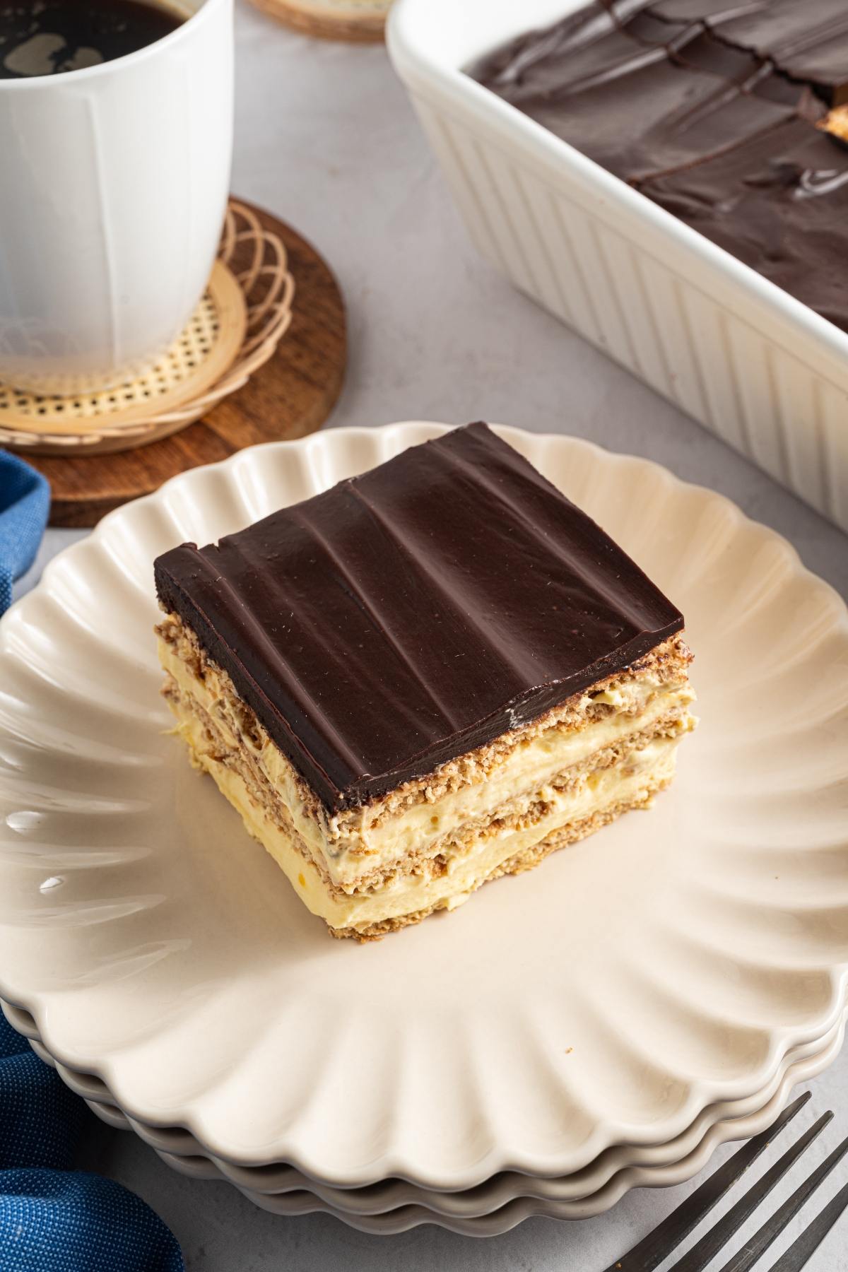 A slice of chocolate-topped layered dessert sits on a white plate next to a cup of coffee.