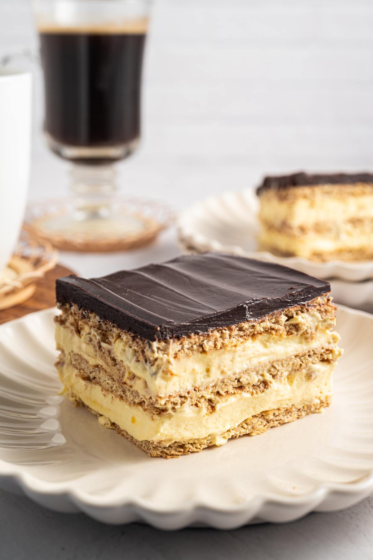 A slice of layered chocolate and cream dessert sits on a plate, with coffee in the background.