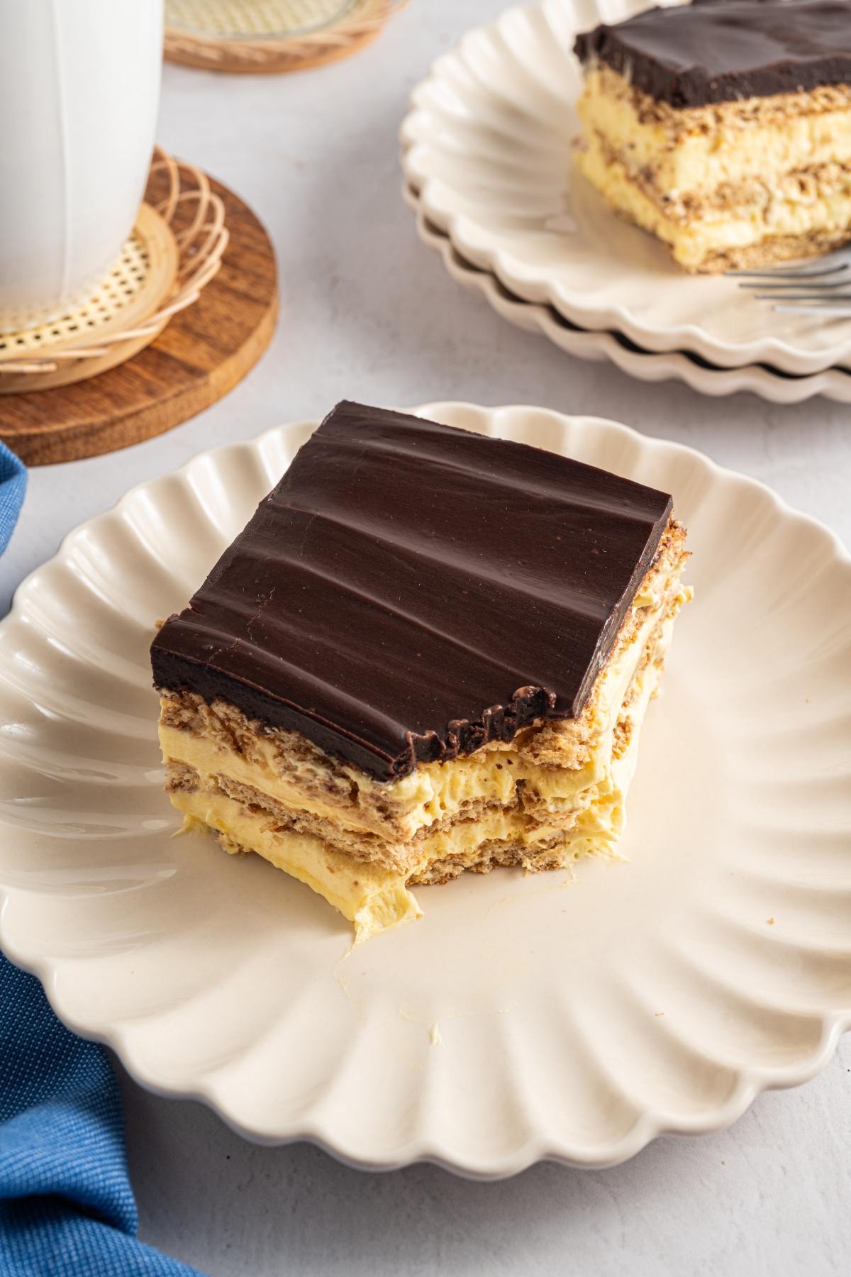 A square slice of chocolate-topped layered dessert sits on a white plate with another slice in the background.
