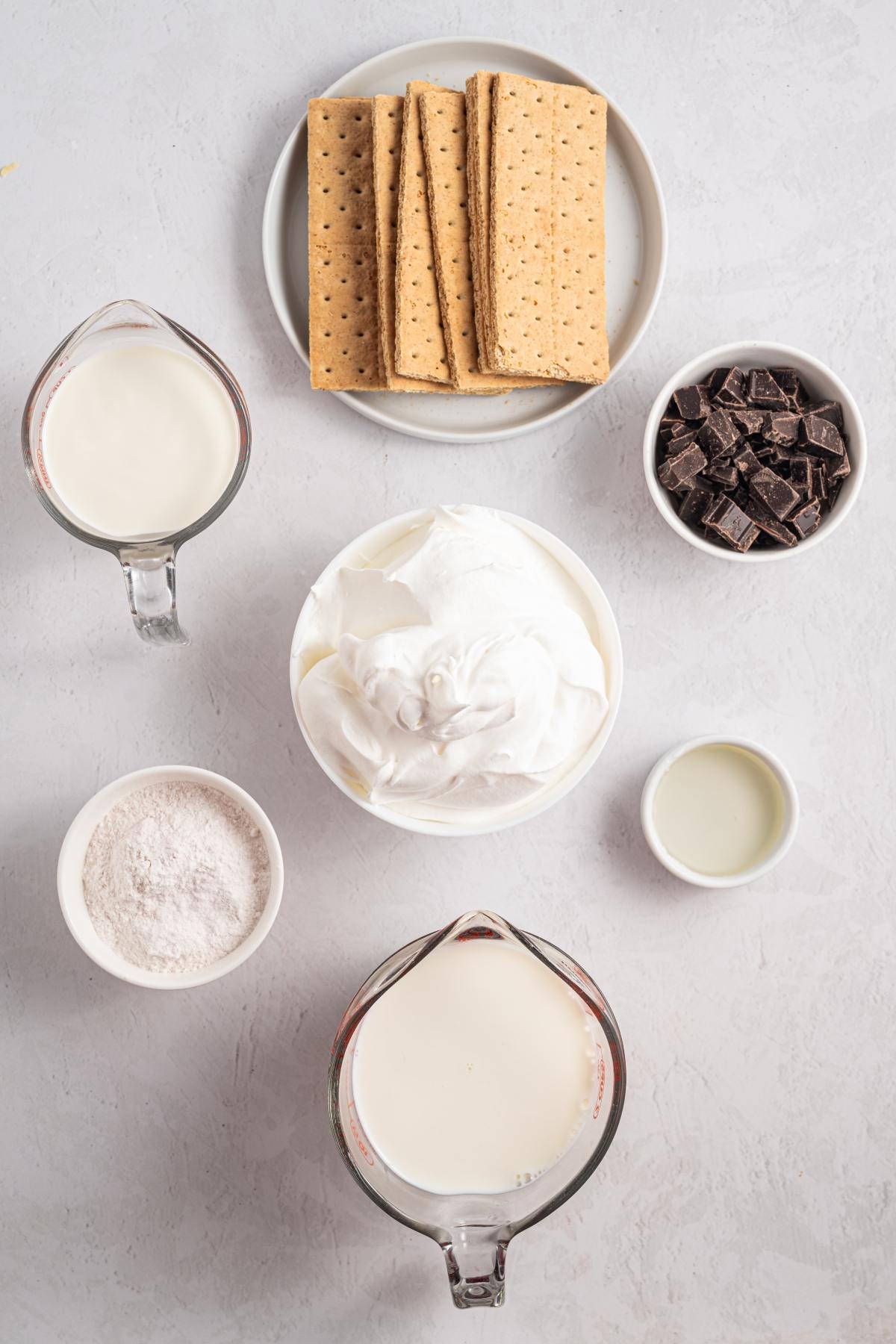 Ingredients for a dessert: graham crackers, chocolate, cream, whipped topping, flour, and milk on a white surface.