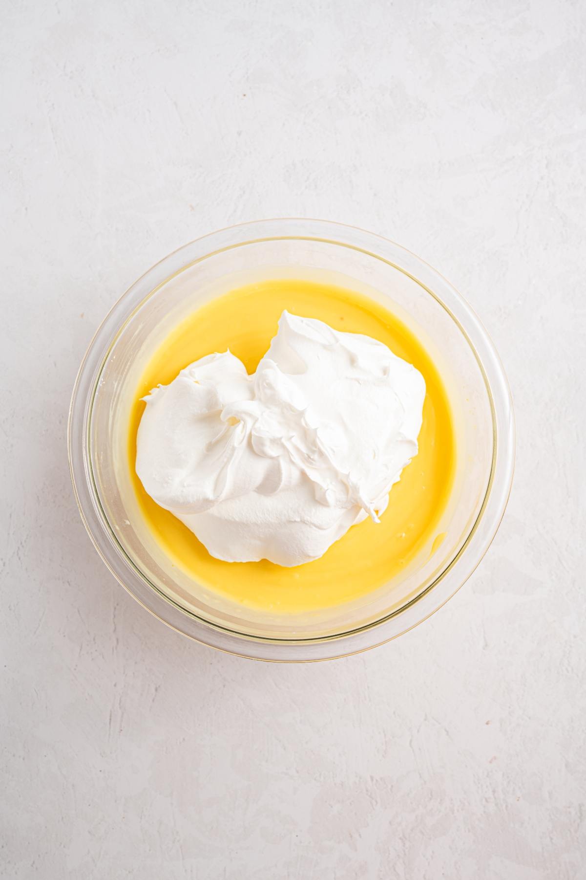 A glass bowl with yellow pudding topped with a large dollop of whipped cream on a white surface.