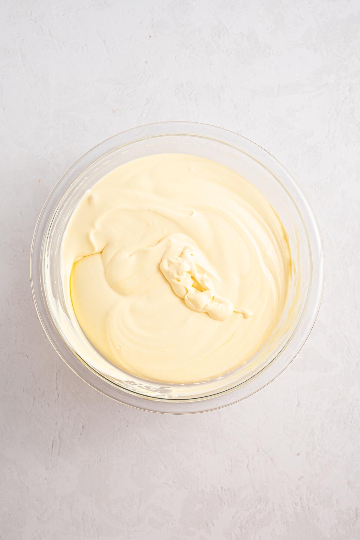 A glass bowl filled with smooth, creamy yellow cheesecake batter on a light surface.