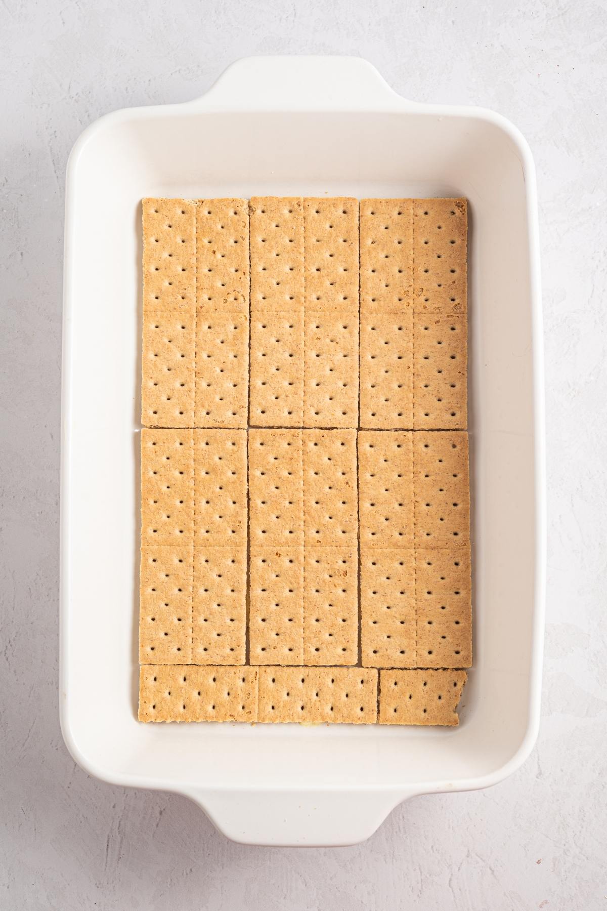 A layer of rectangular graham crackers neatly arranged in a white rectangular baking dish.