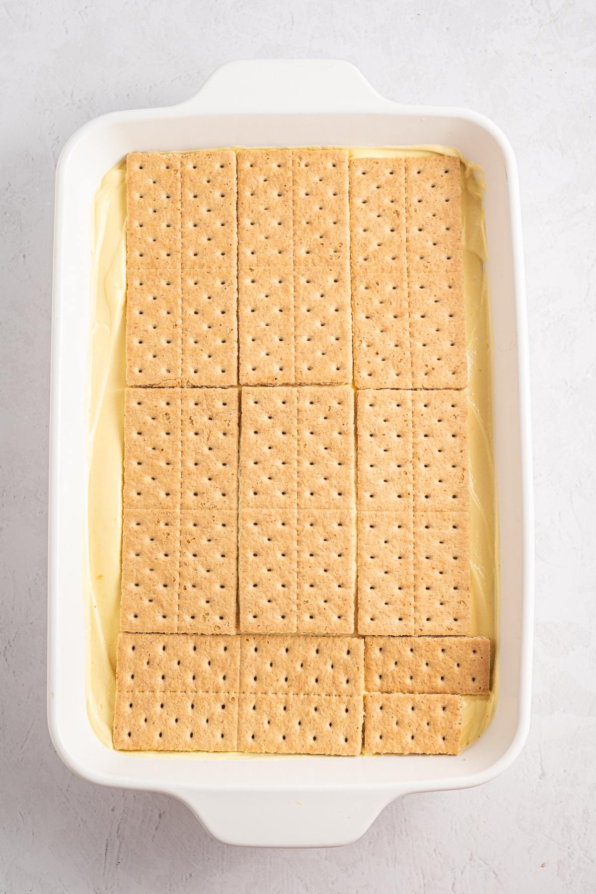Rectangular graham crackers layered over a creamy yellow filling in a white baking dish.