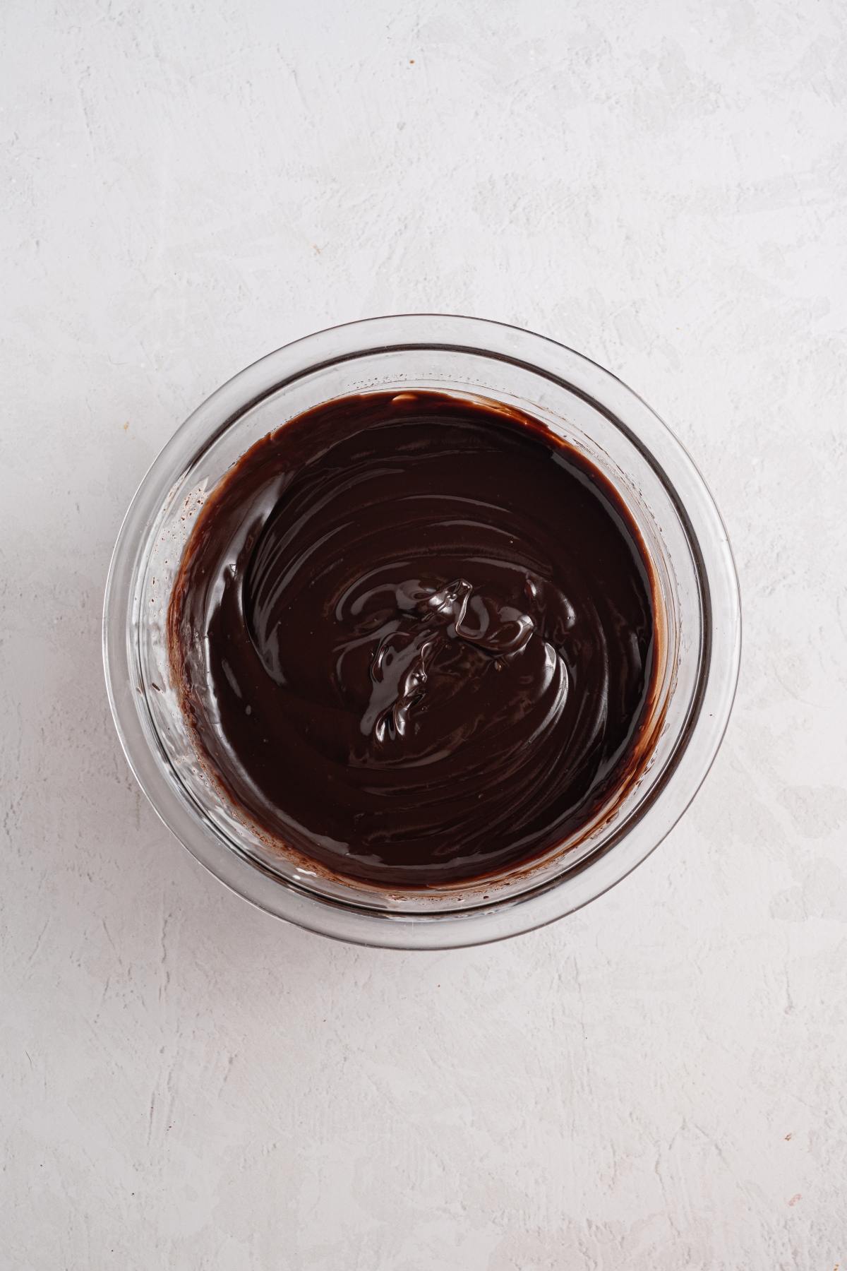 A glass bowl filled with smooth, shiny melted chocolate on a light textured surface.