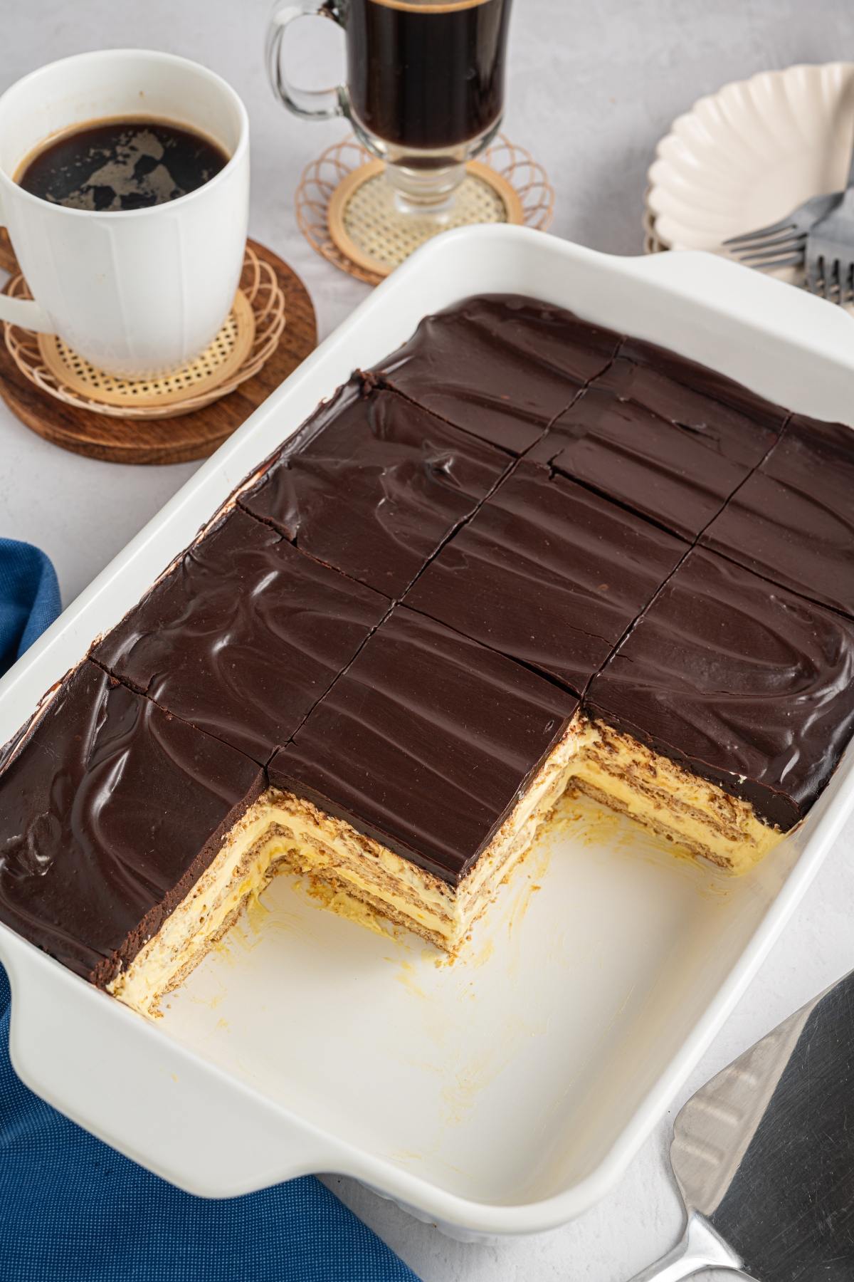 A rectangular chocolate-covered layered dessert in a white dish, with several pieces cut and served.