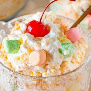 A glass bowl of creamy Frog Eye Salad is topped with colorful mini marshmallows and a red cherry.