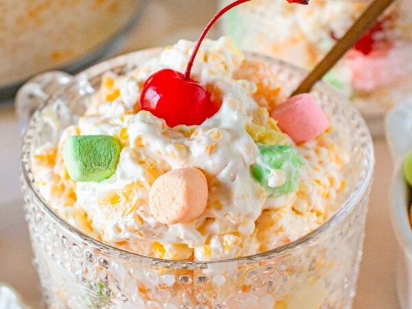 A glass bowl of creamy Frog Eye Salad is topped with colorful mini marshmallows and a red cherry.