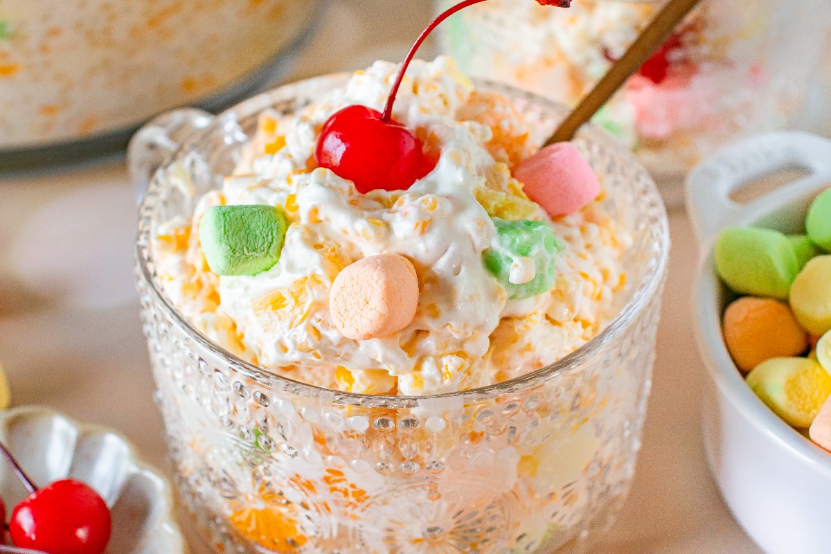 A glass bowl of creamy Frog Eye Salad features colorful mini marshmallows, fruit, and a cherry on top.