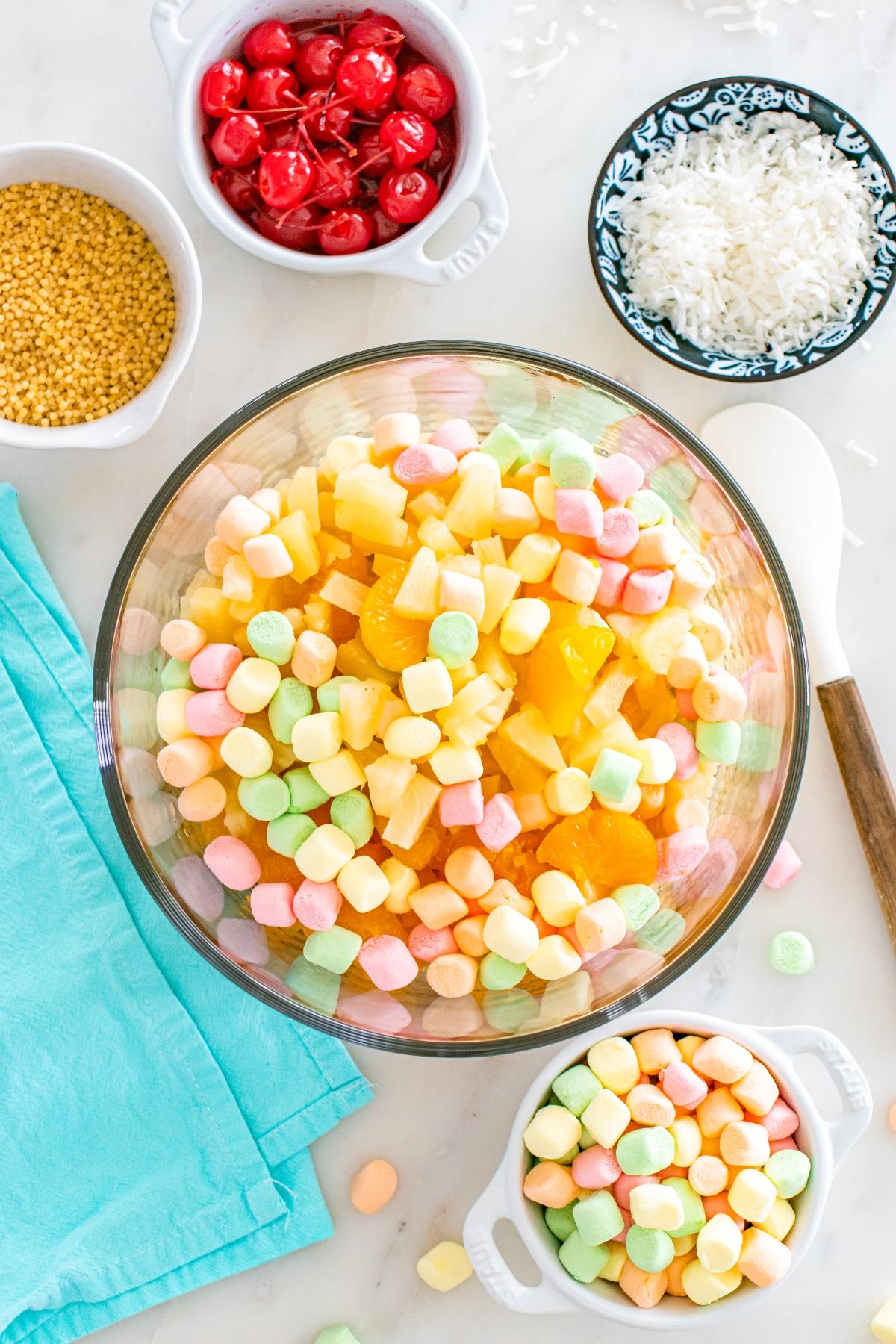 A bowl of fruit salad with colorful marshmallows, surrounded by cherries, coconut, and sprinkles.