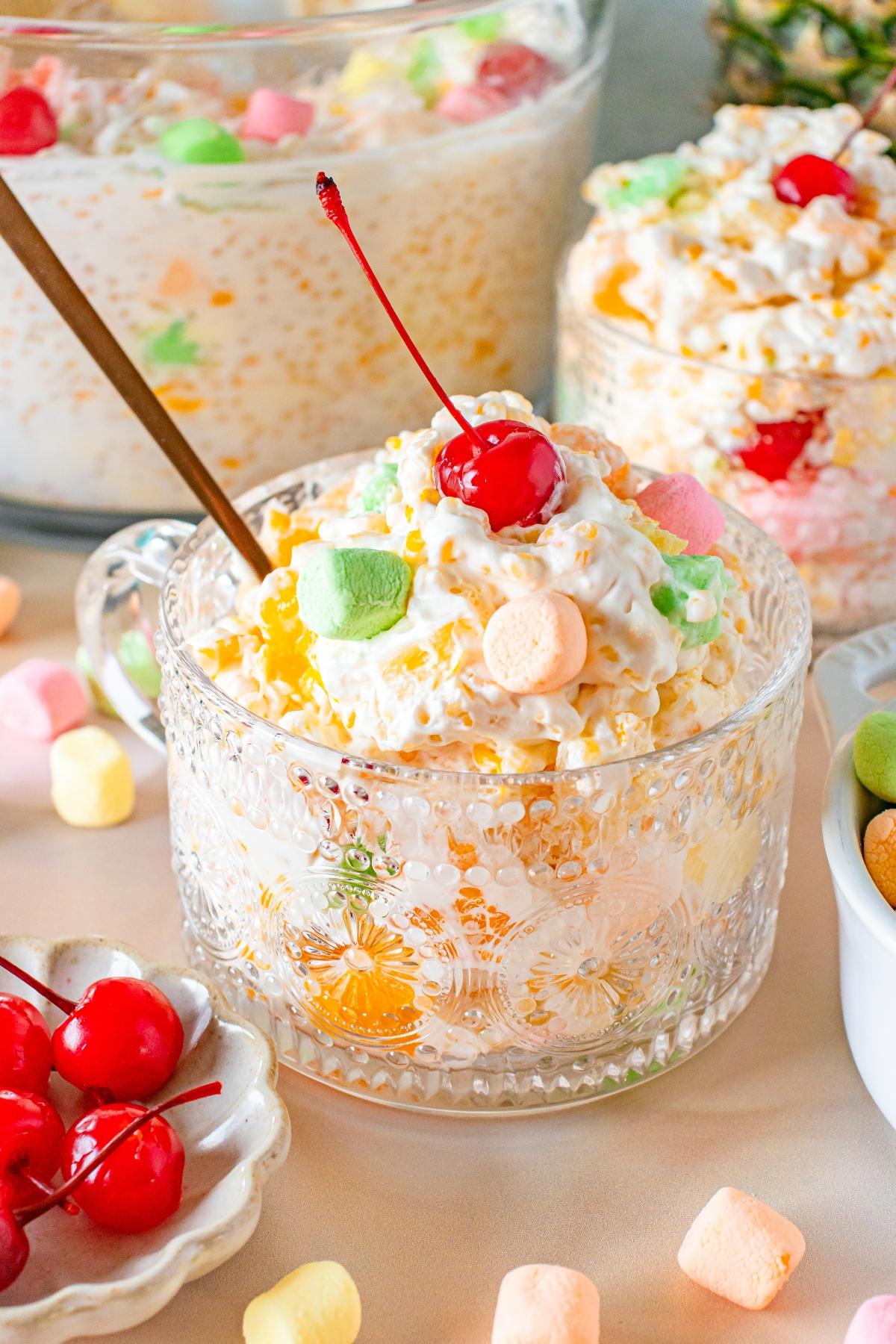 Glass cup of creamy frog eye fruit salad with mini marshmallows, topped with a cherry, on a table with more marshmallows.