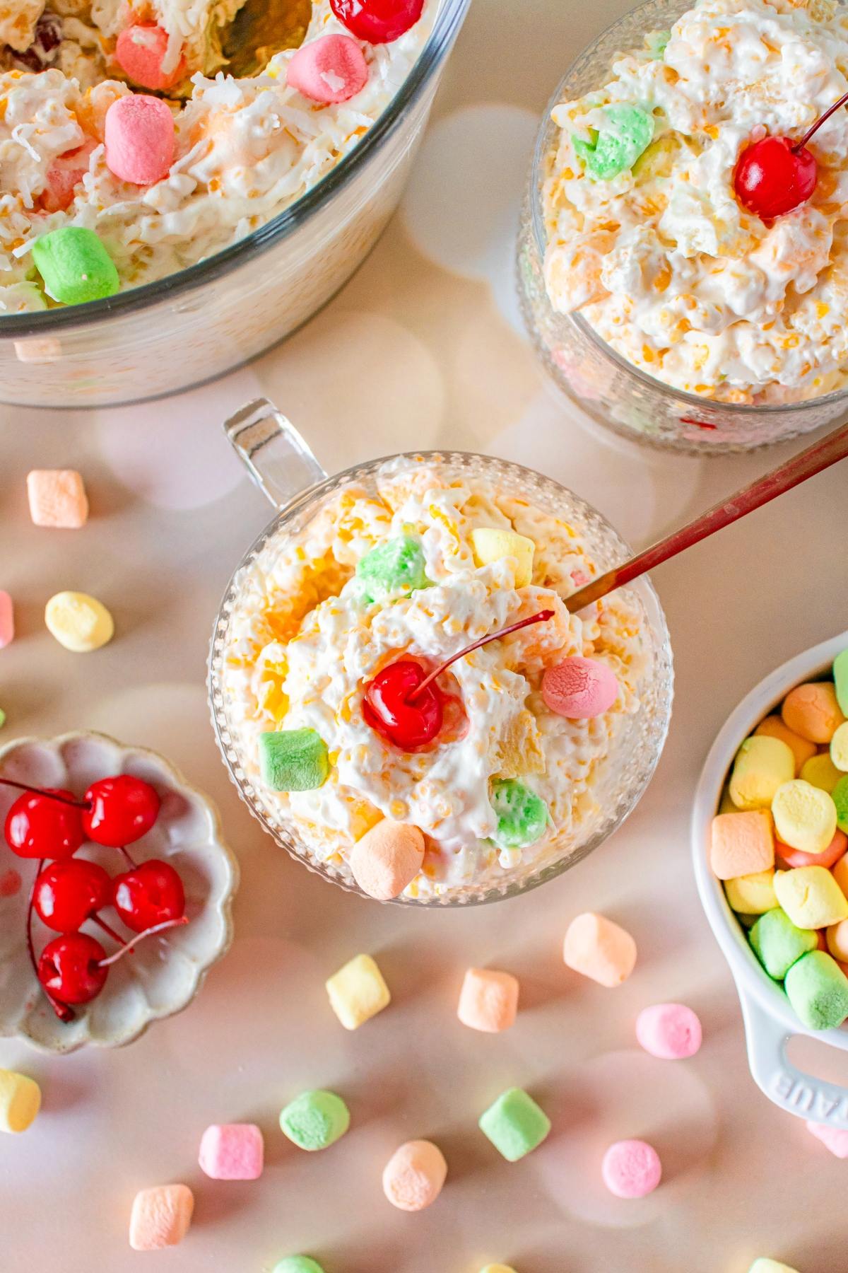 Glass cups filled with creamy frog eye fruit salad, topped with cherries and colorful mini marshmallows on a light table.