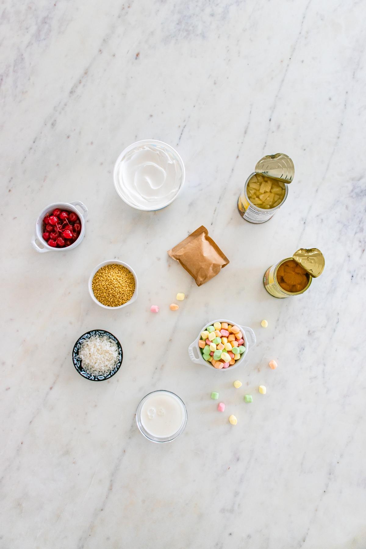 Assorted dessert ingredients in bowls and cans arranged on a white marble surface, including fruit and marshmallows.