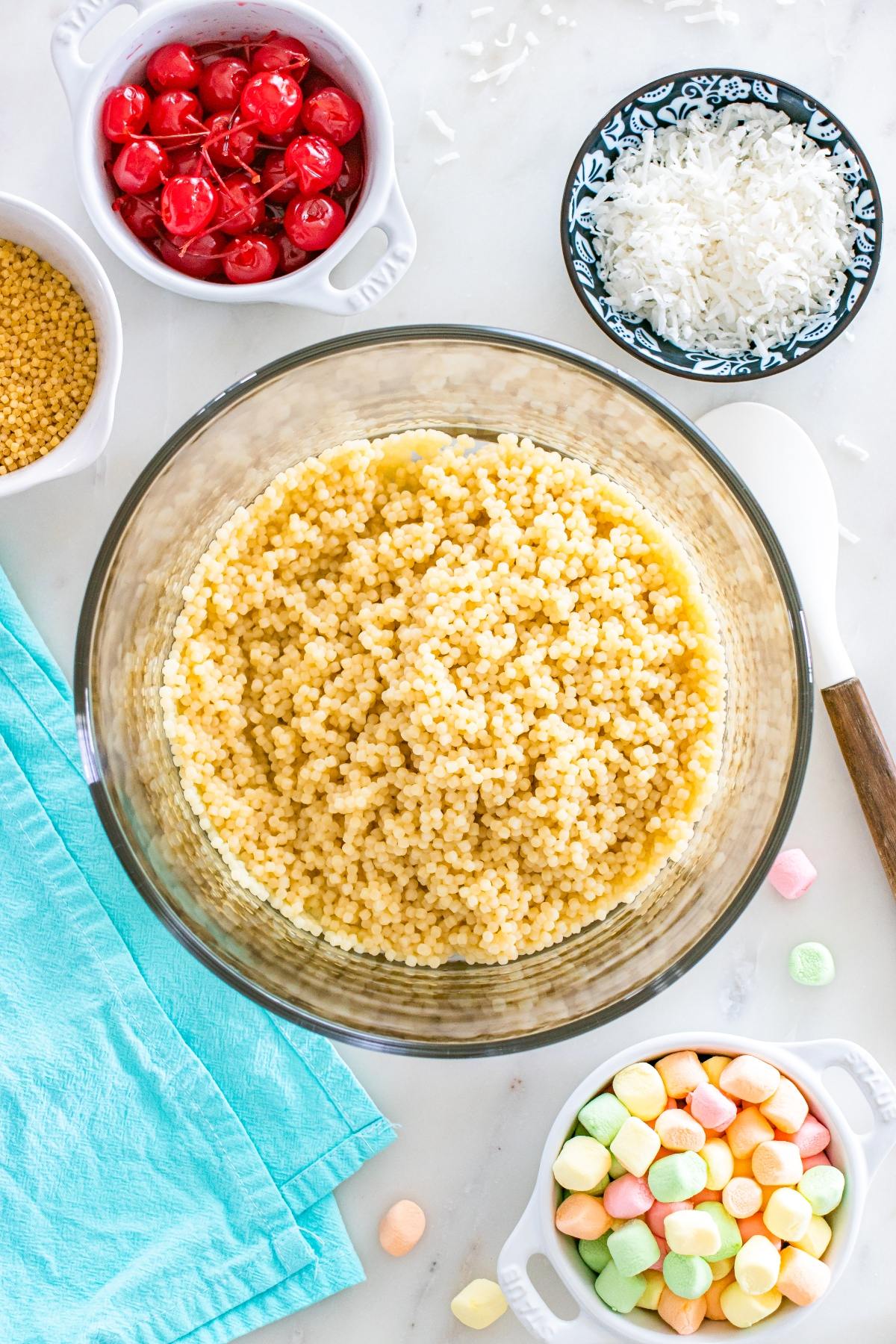 A bowl of cooked acini di pepe pasta surrounded by cherries, coconut, nonpareils, and mini marshmallows.