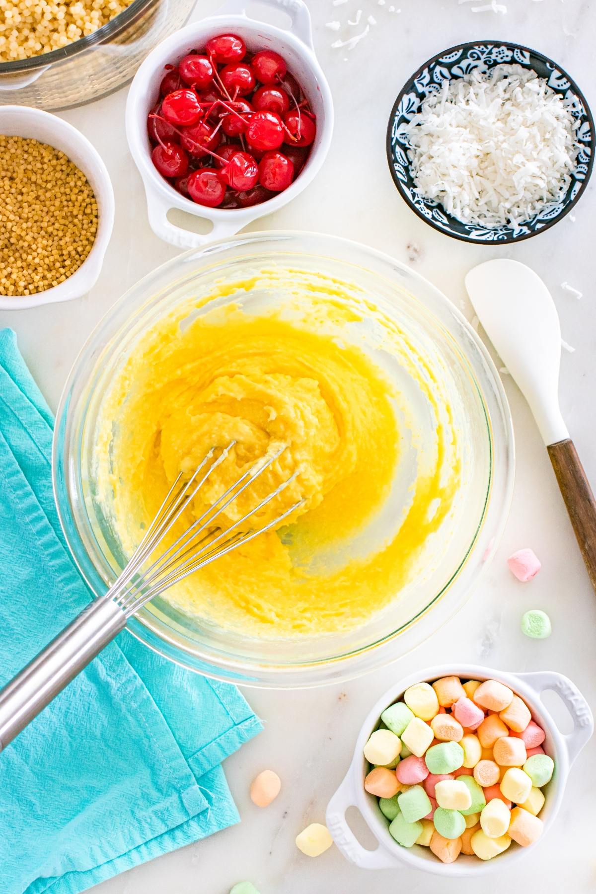 Bowl of yellow cake batter with whisk, surrounded by cherries, coconut, crisp cereal, and colorful marshmallows.