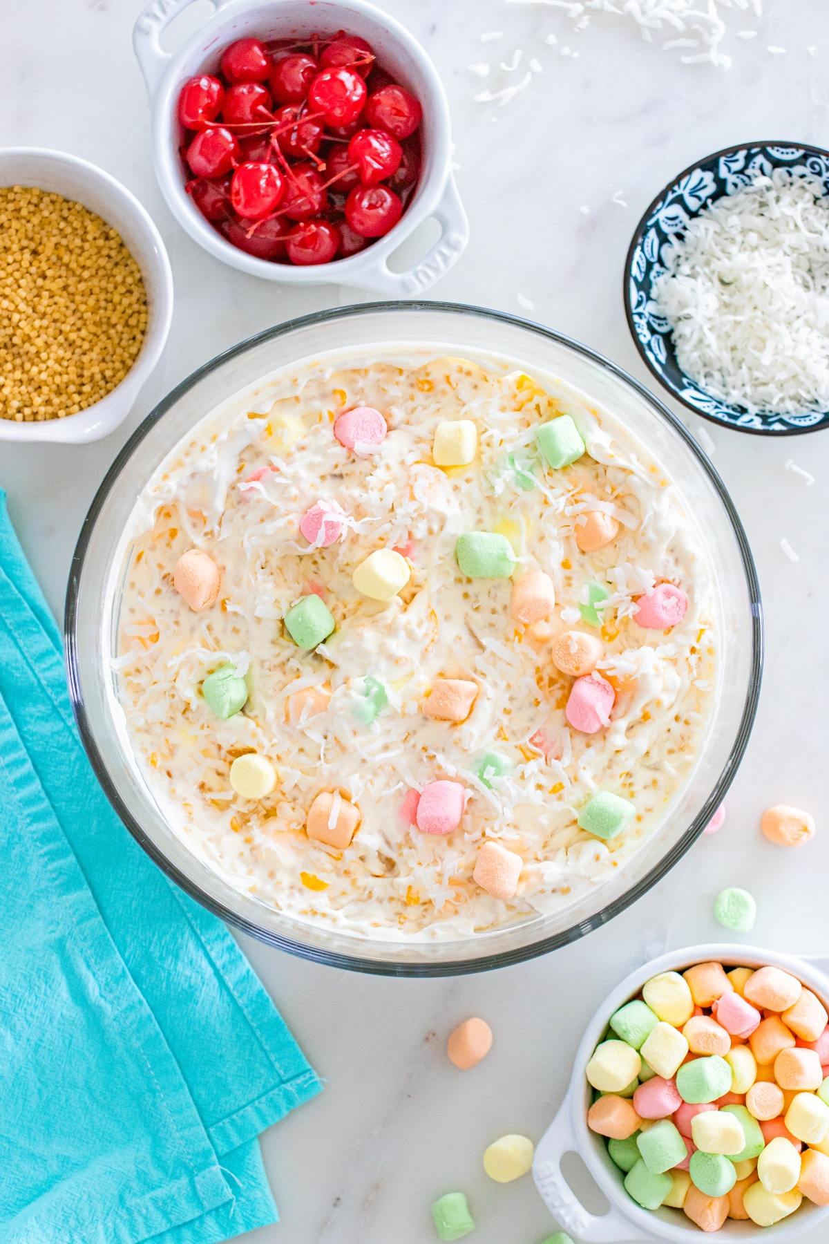 A bowl of creamy fruit salad topped with colorful mini marshmallows, surrounded by ingredients in small bowls.