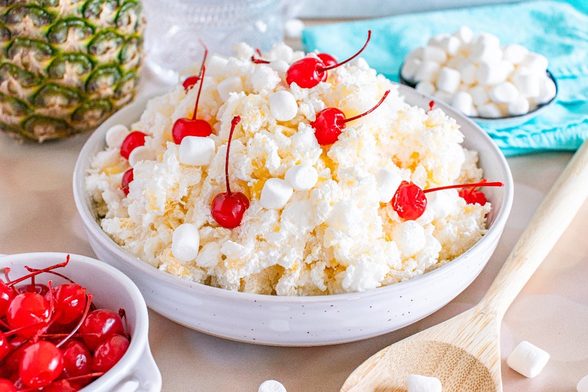 A bowl of pineapple fluff salad, reminiscent of classic glorified rice, is topped with maraschino cherries and mini marshmallows on a table.