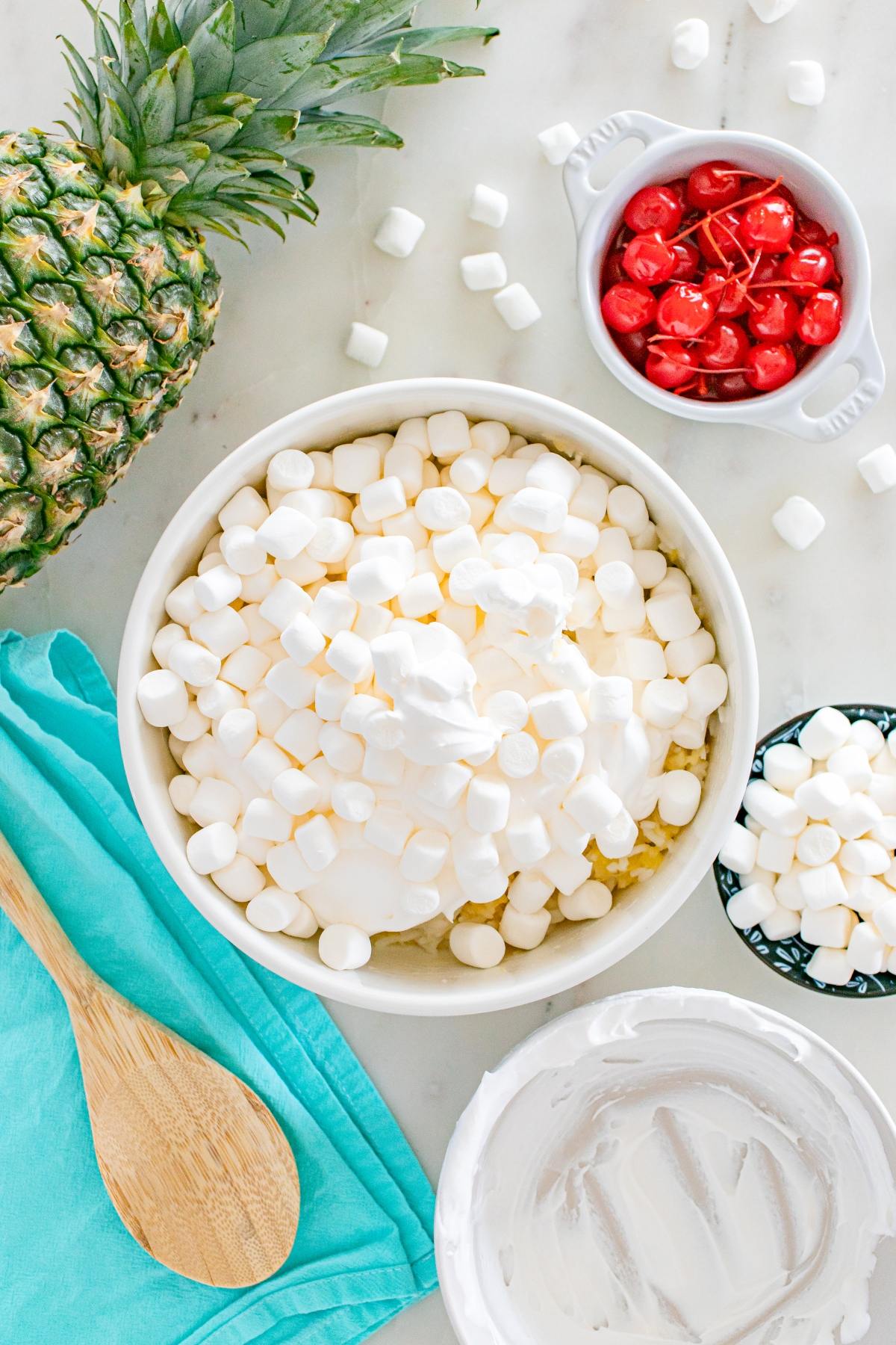A bowl of marshmallows and whipped topping, with pineapple, cherries, and a wooden spoon on a counter.
