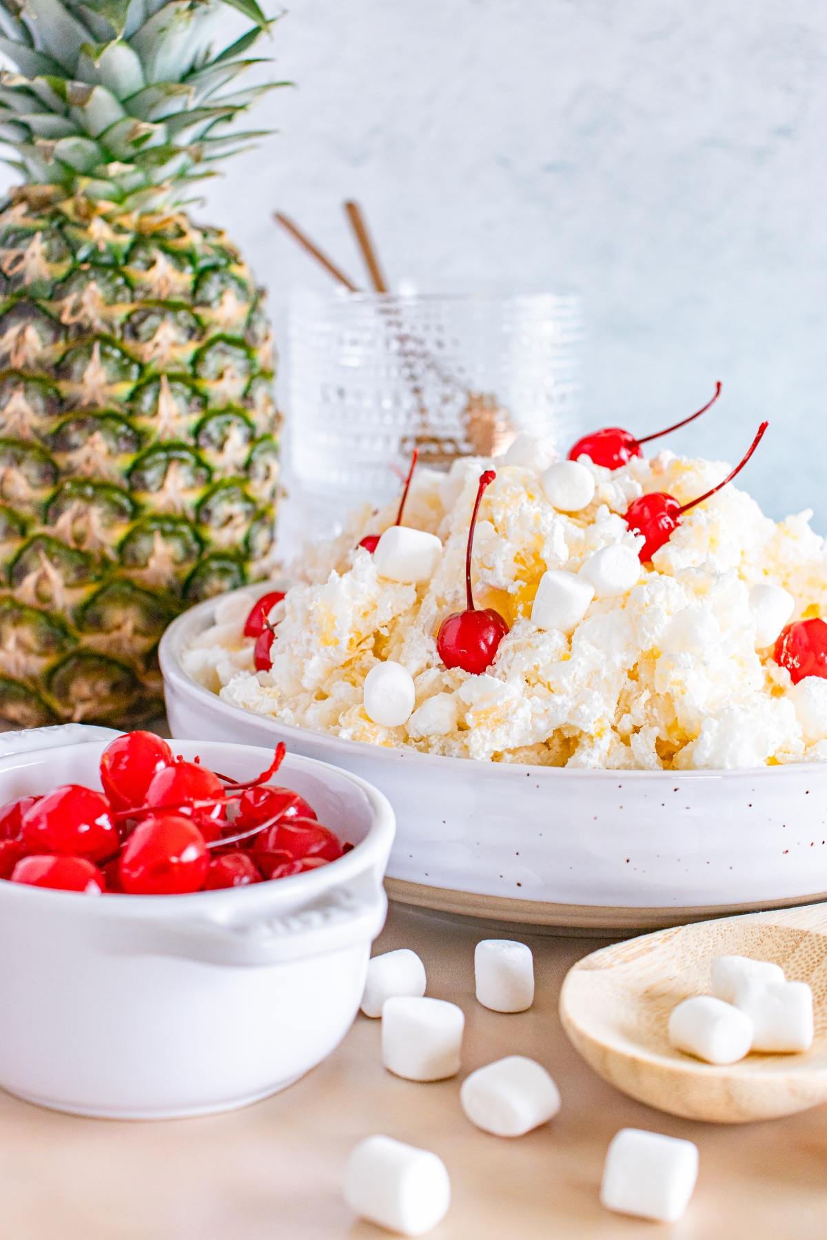 A bowl of glorified rice  ambrosia salad with cherries and marshmallows, next to a fresh pineapple and a dish of cherries.