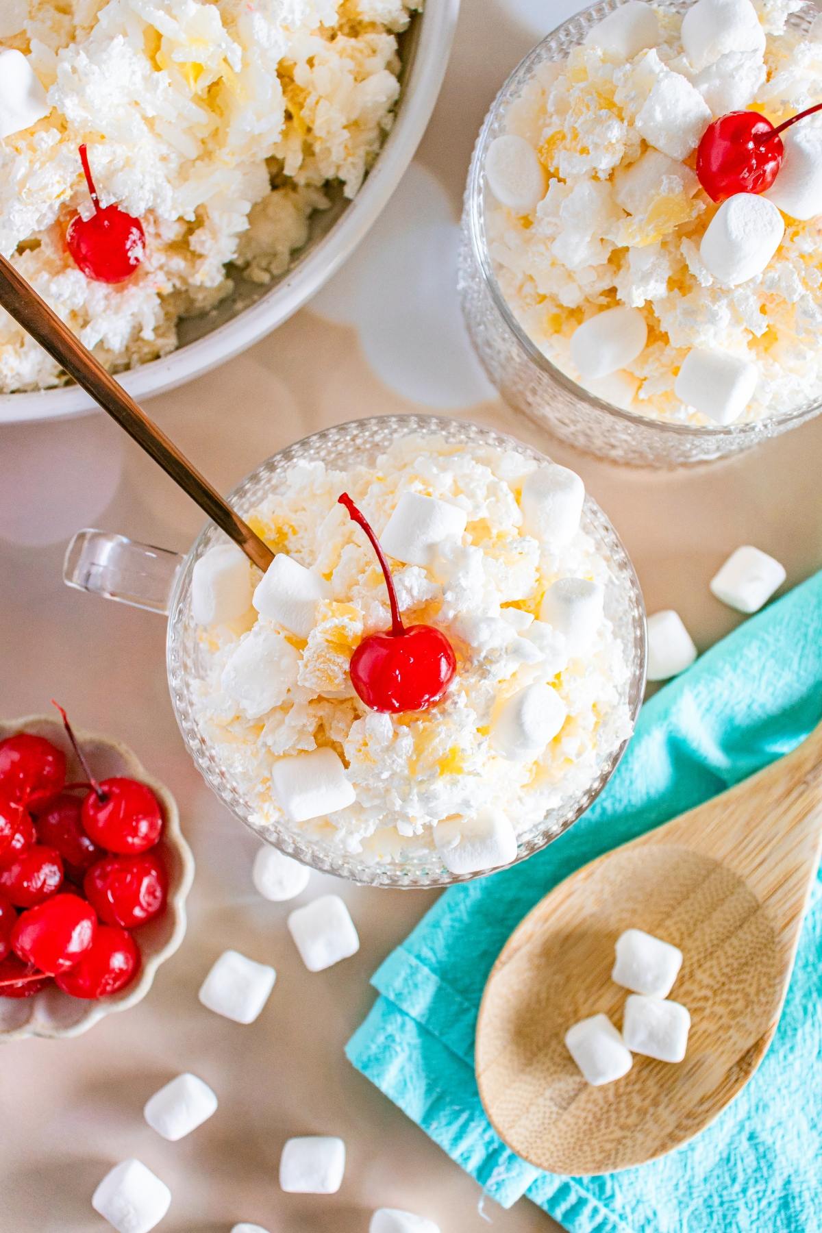 Two glass bowls of glorified rice  ambrosia salad topped with mini marshmallows and cherries, with extra cherries and marshmallows nearby.