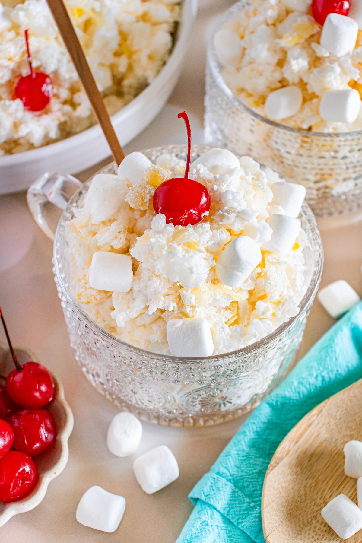 A glass bowl of glorified rice creamy dessert topped with mini marshmallows and a cherry, with more cherries and marshmallows nearby.