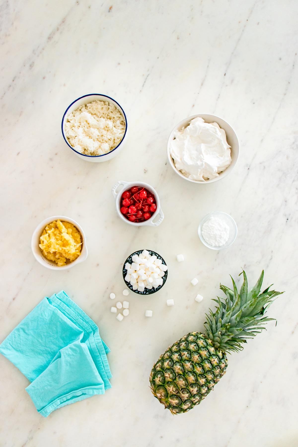Ingredients for a dessert, including pineapple, whipped cream, marshmallows, fruit, and blue napkins on a marble surface.