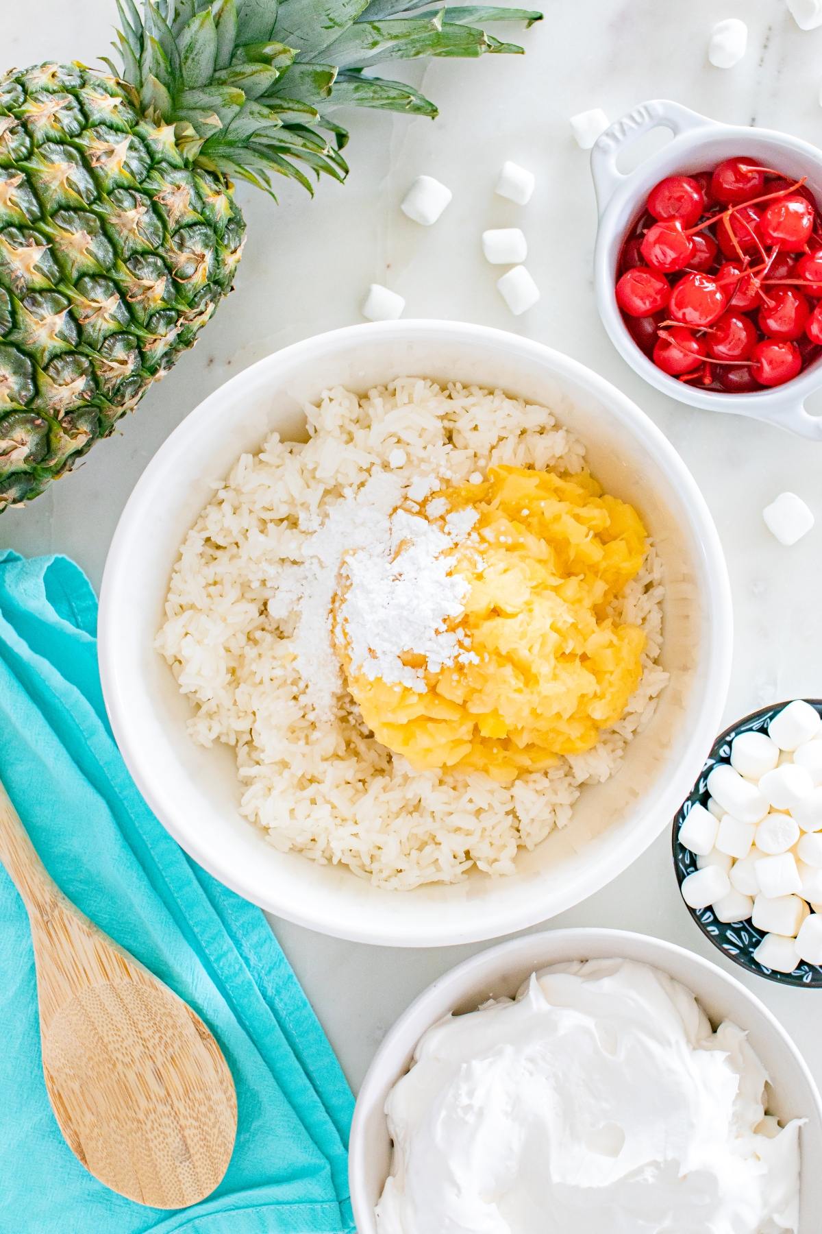 Bowl with rice, pineapple, sugar, and marshmallows, surrounded by cherries, whipped topping, and a whole pineapple.