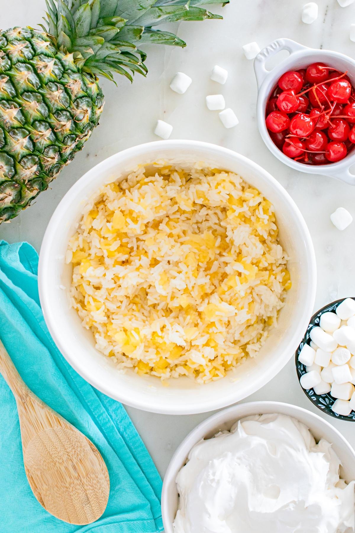 Bowl of pineapple rice surrounded by pineapple, cherries, marshmallows, whipped topping, and a wooden spoon.