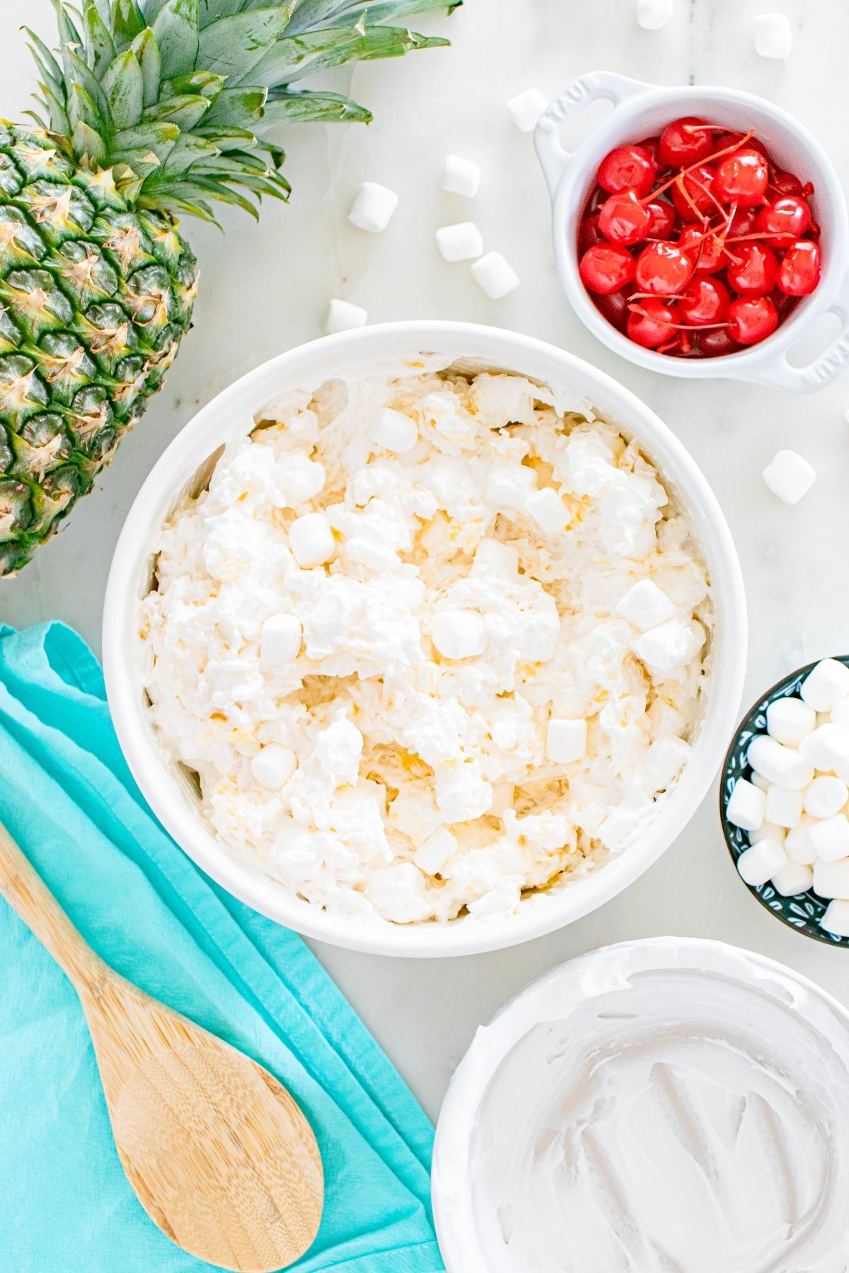 Bowl of creamy fruit salad with marshmallows, surrounded by pineapple, cherries, and a wooden spoon.