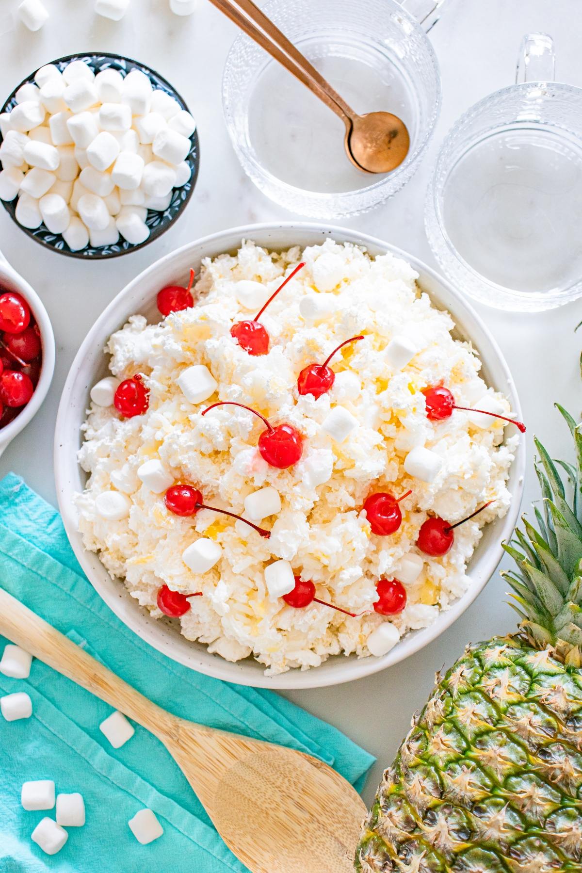 A bowl of glorified rice  ambrosia salad with cherries and marshmallows, surrounded by pineapple, marshmallows, and glass cups.