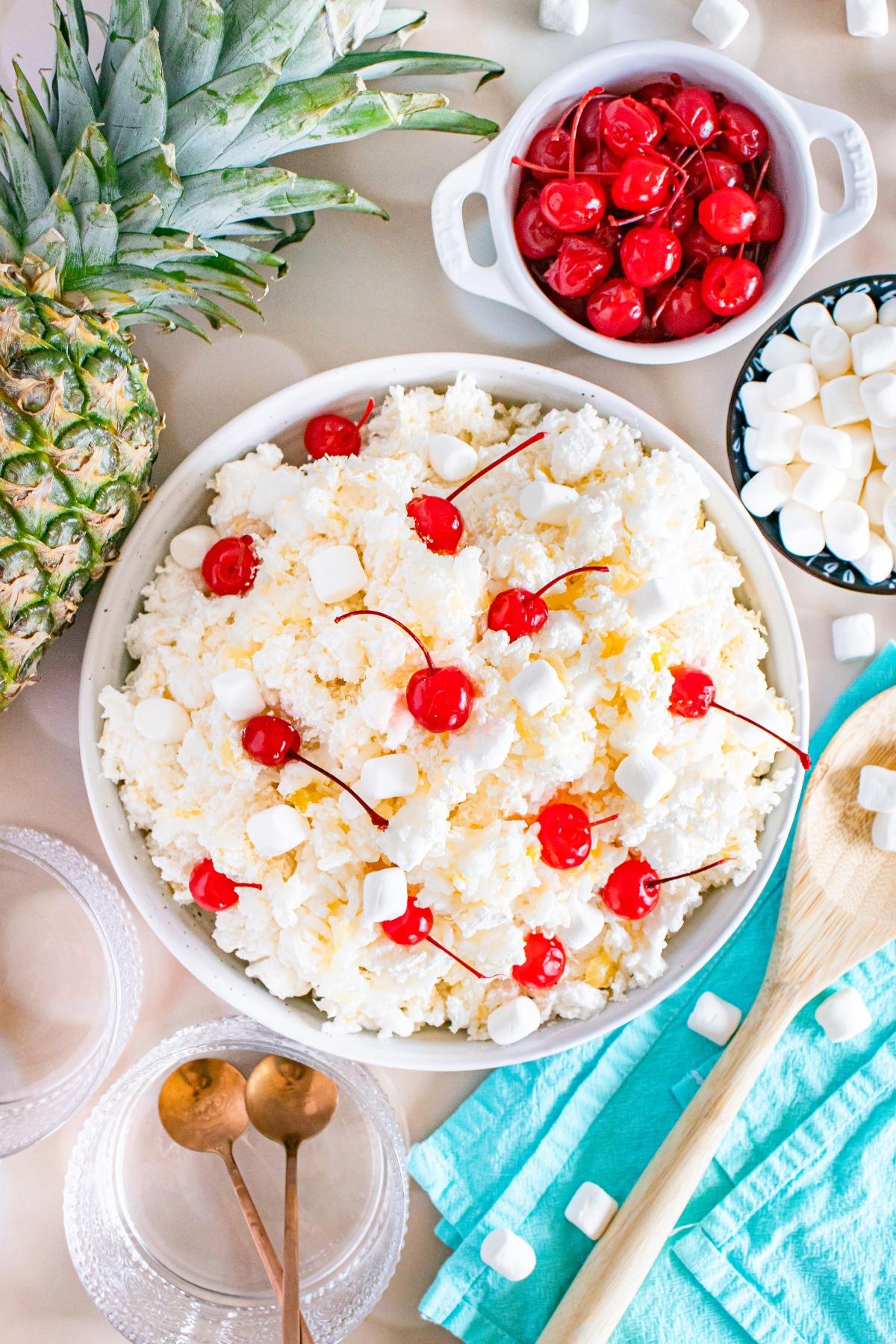 A bowl of glorified rice creamy dessert topped with cherries and mini marshmallows, surrounded by pineapple and ingredients.