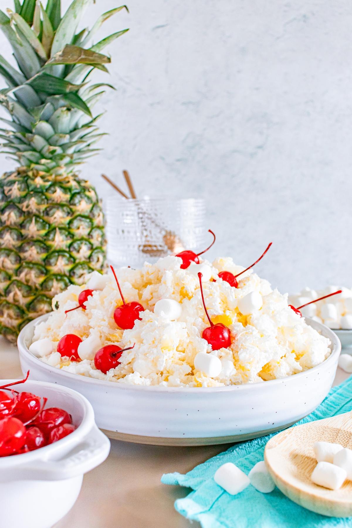 A bowl of glorified rice  fluffy white salad topped with cherries and mini marshmallows, with a whole pineapple nearby.