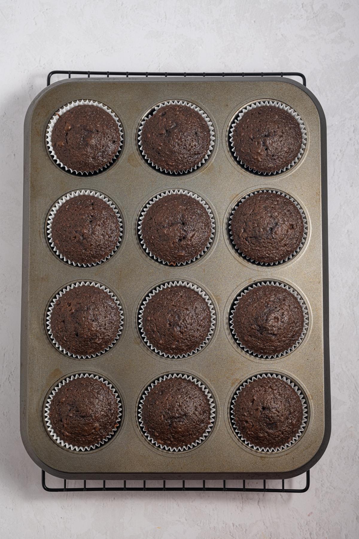 A muffin tin with twelve baked chocolate cupcakes in silver paper liners on a cooling rack.
