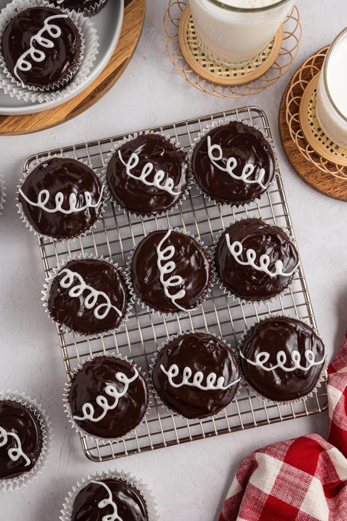 Chocolate homemade hostess cupcakes with shiny frosting and white icing swirls, on a cooling rack next to glasses of milk.