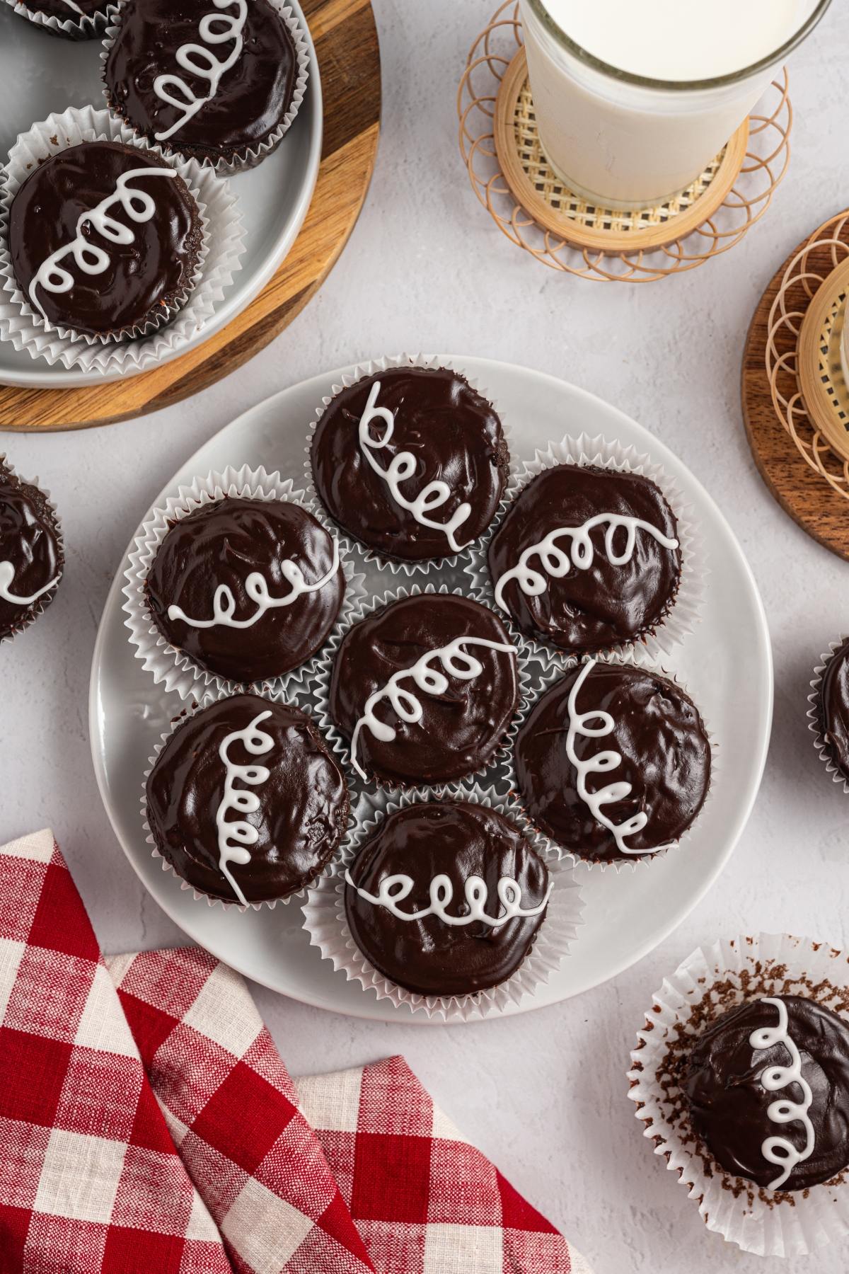 Chocolate homemade hostess cupcakes with white icing swirls on a plate, next to a glass of milk and a red checkered napkin.