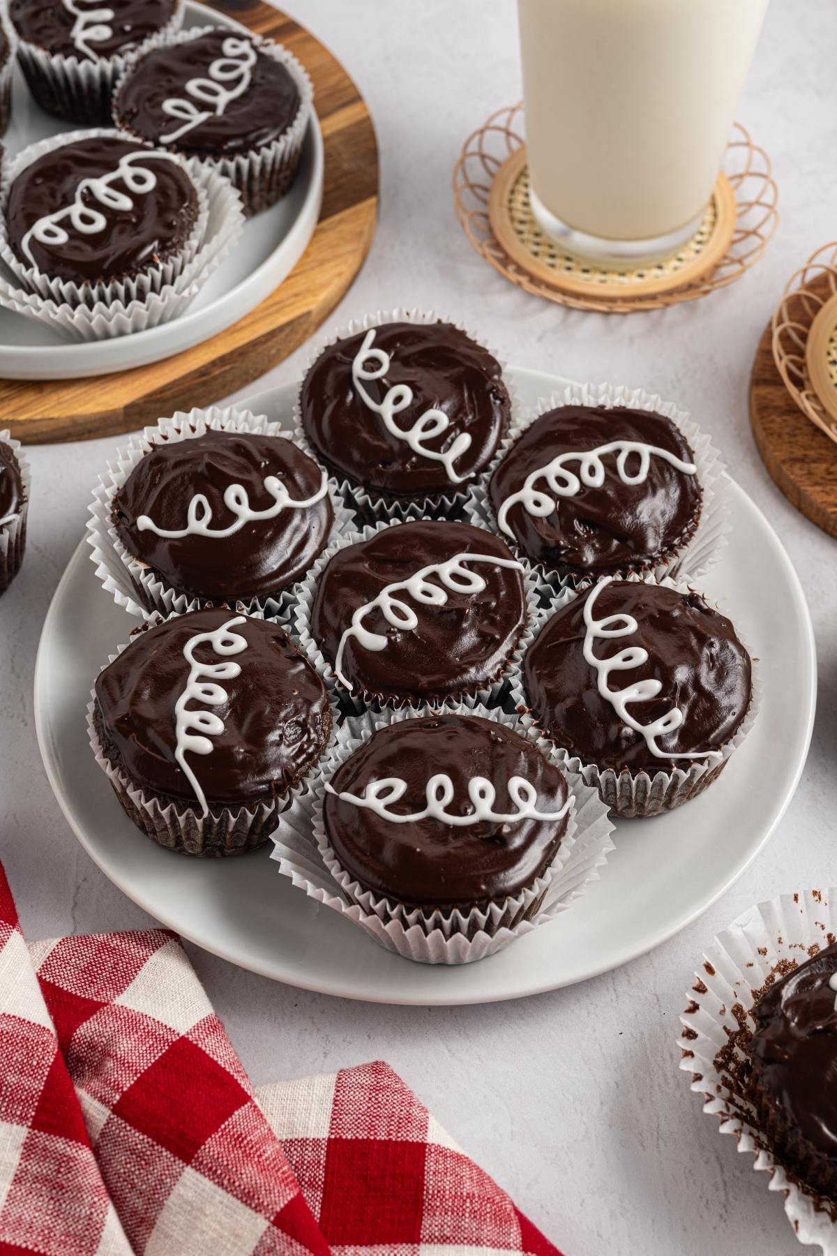 Chocolate homemade hostess cupcakes with white icing swirls on a plate, with a glass of milk and a red checked napkin nearby.