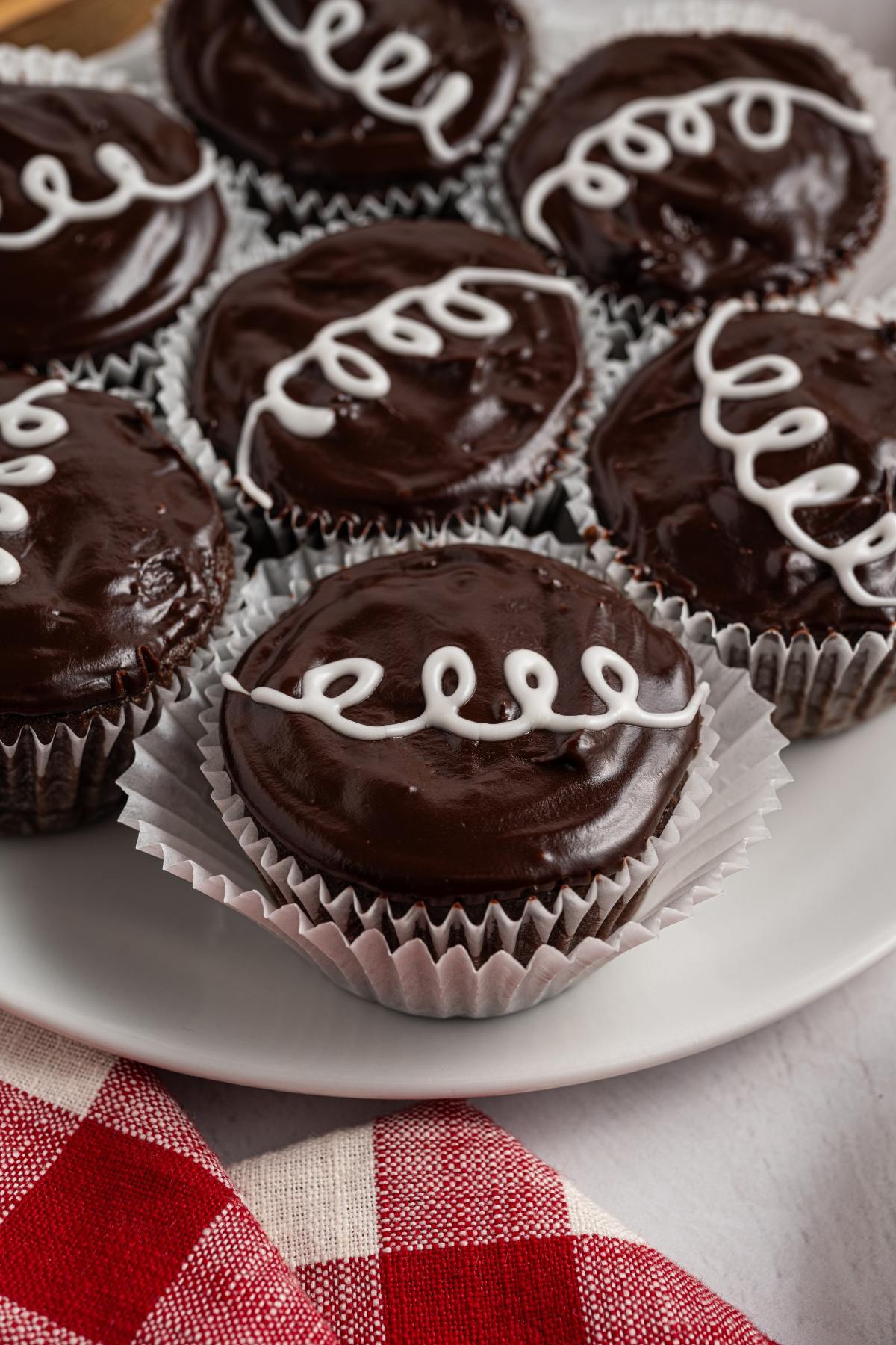 Chocolate homemade hostess cupcakes with glossy frosting and white icing swirls on a white plate, next to a red checked cloth.